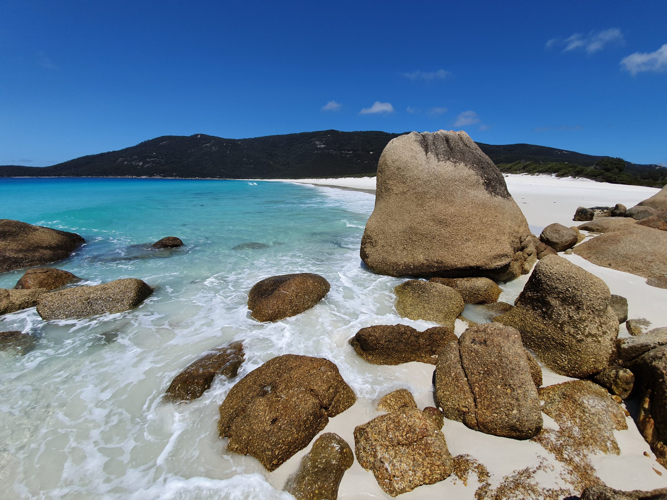 Large rocks on a white sandy beach with gentle waves from clear ocean, and grassy headland in the distance.