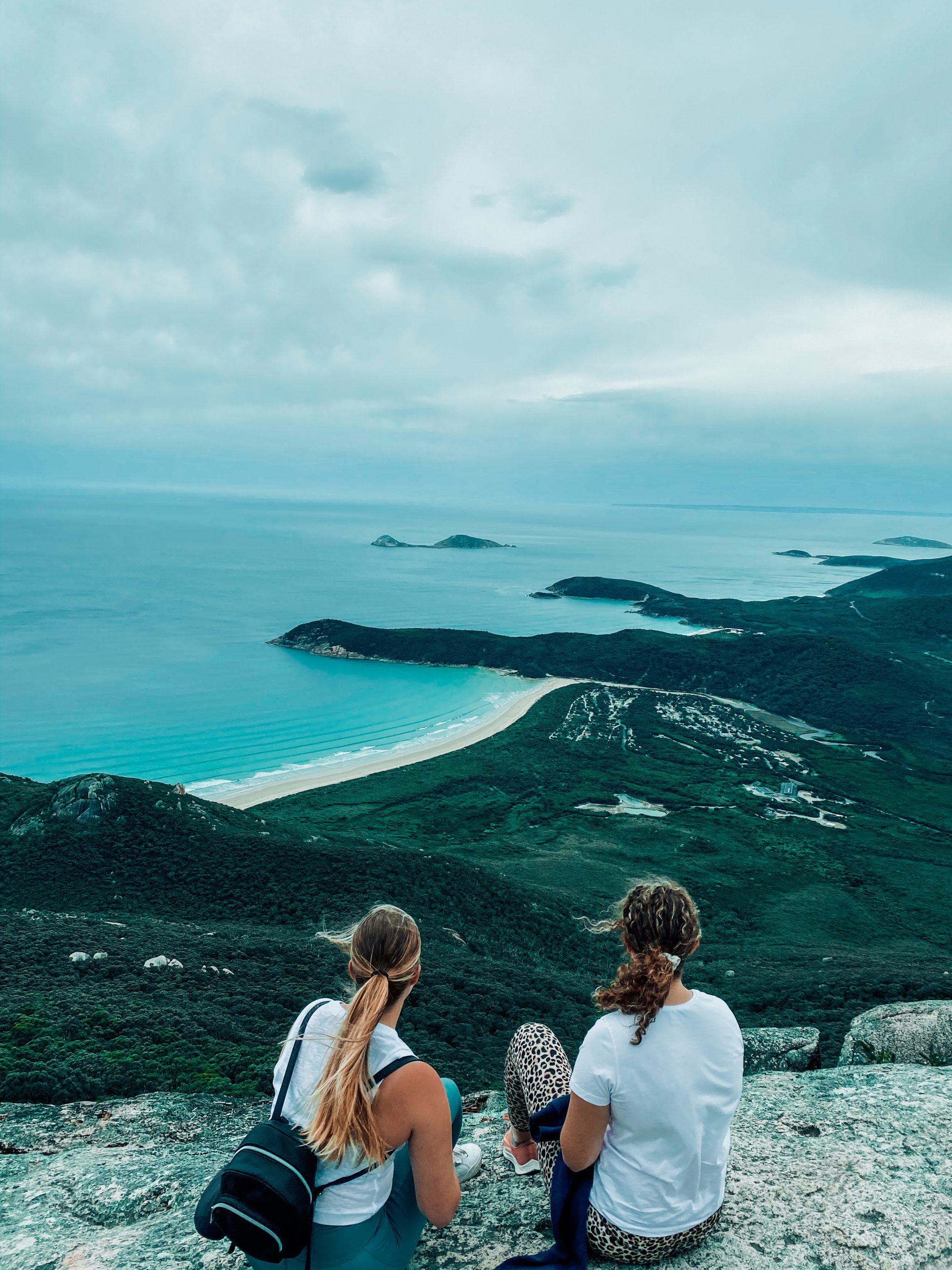 Two people sat on a rock overlooking a coastline with beaches, grassy headlands, blue ocean and islands in the distance.