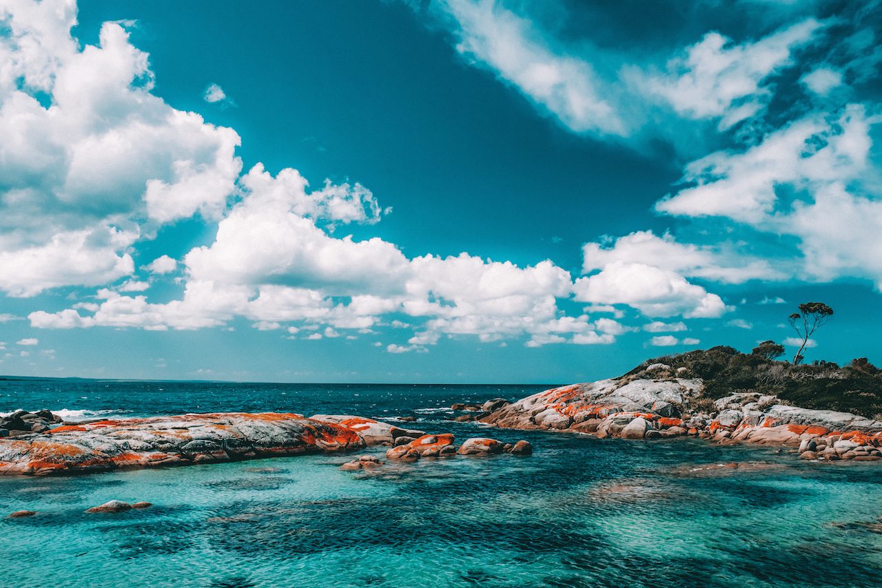 A bay with orange rocks and clear turquoise water against a blue sky with dotted white clouds.