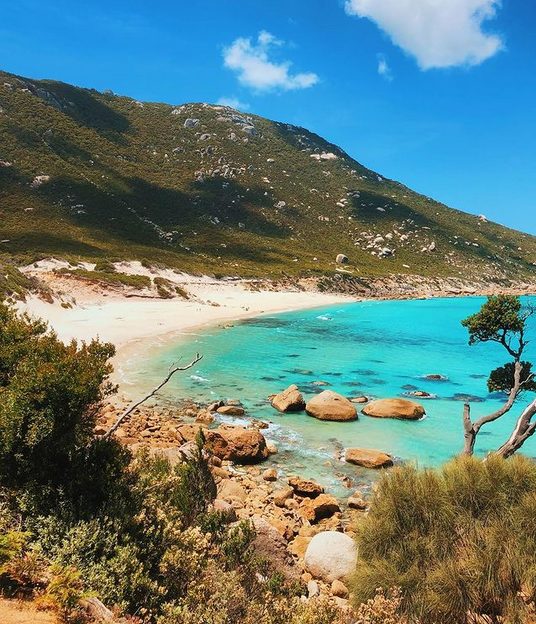 A small bay with turquoise blue ocean, small rocks dotted in the water, white sand and steep bushland behind it.