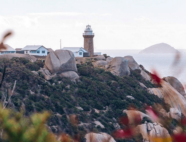 A lighthouse and some small white buildings on the top of a rocky cliff by the ocean.