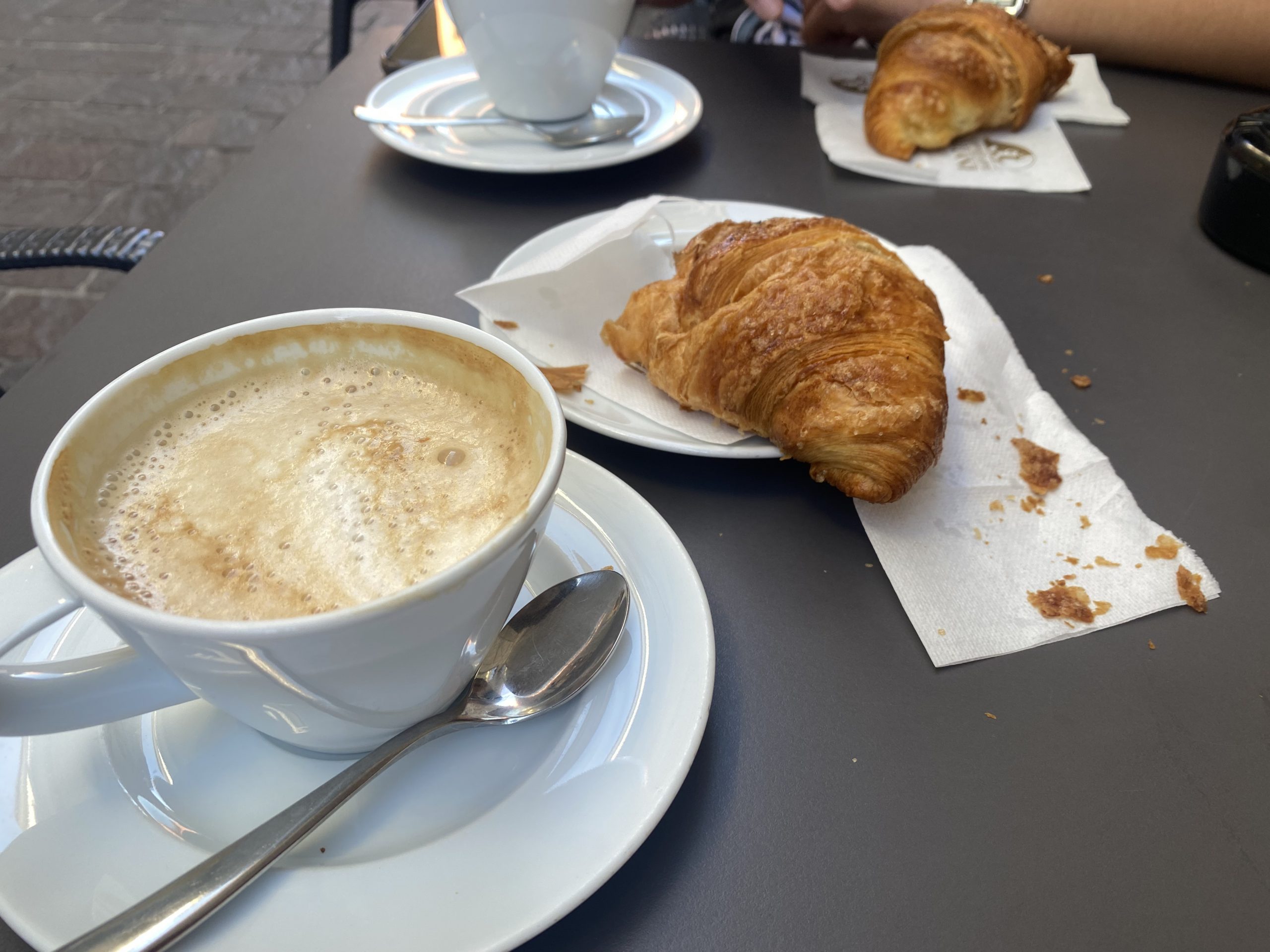 Frothy coffees in white mugs and croissants on plates on a grey table.