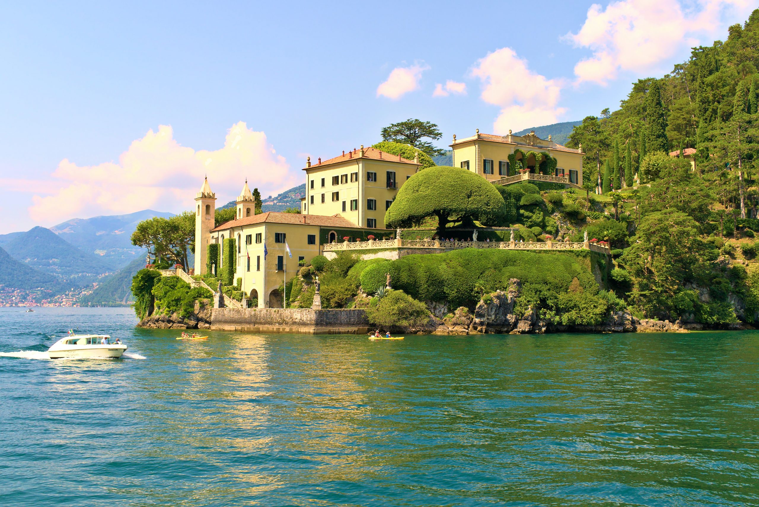 A yellow italian villa with an orange terracotta roof and manicured gardens set above a blue lake with a speed boat driving past in the foreground.