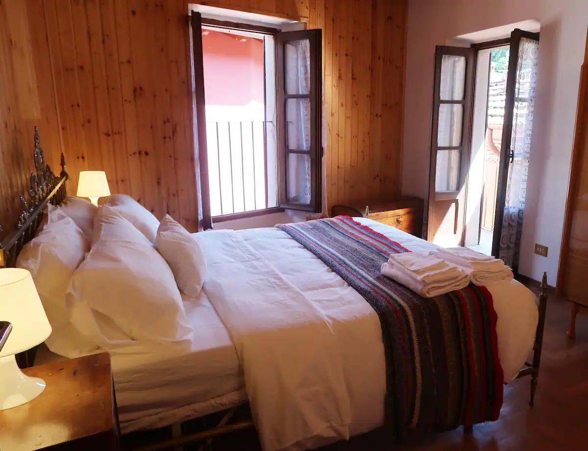 a bedroom with a double bed, wooden panelling, dressers, lamps and open french windows.