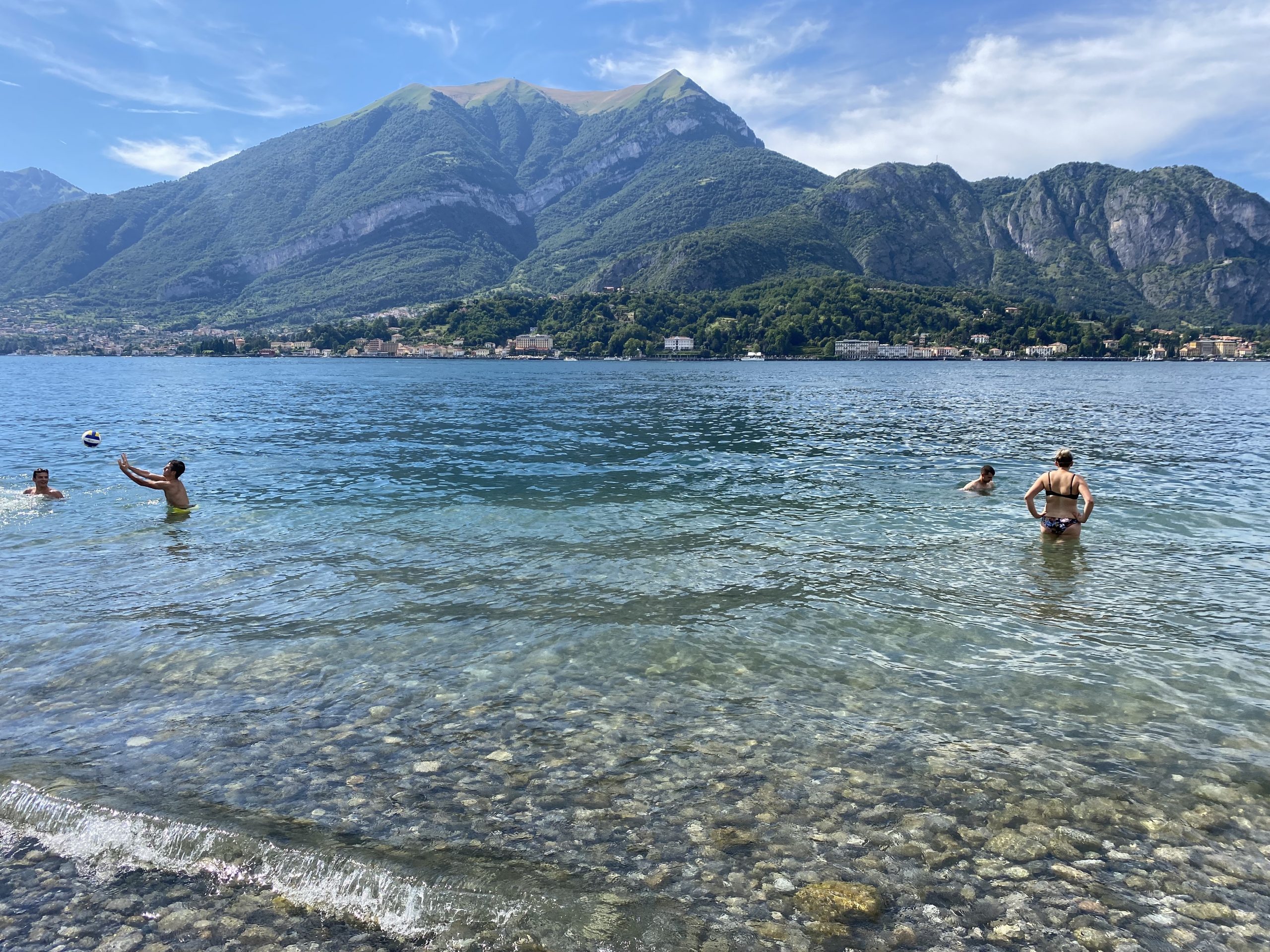 A pebble beach with four people swimming in the blue lake and mountains and towns in the background.