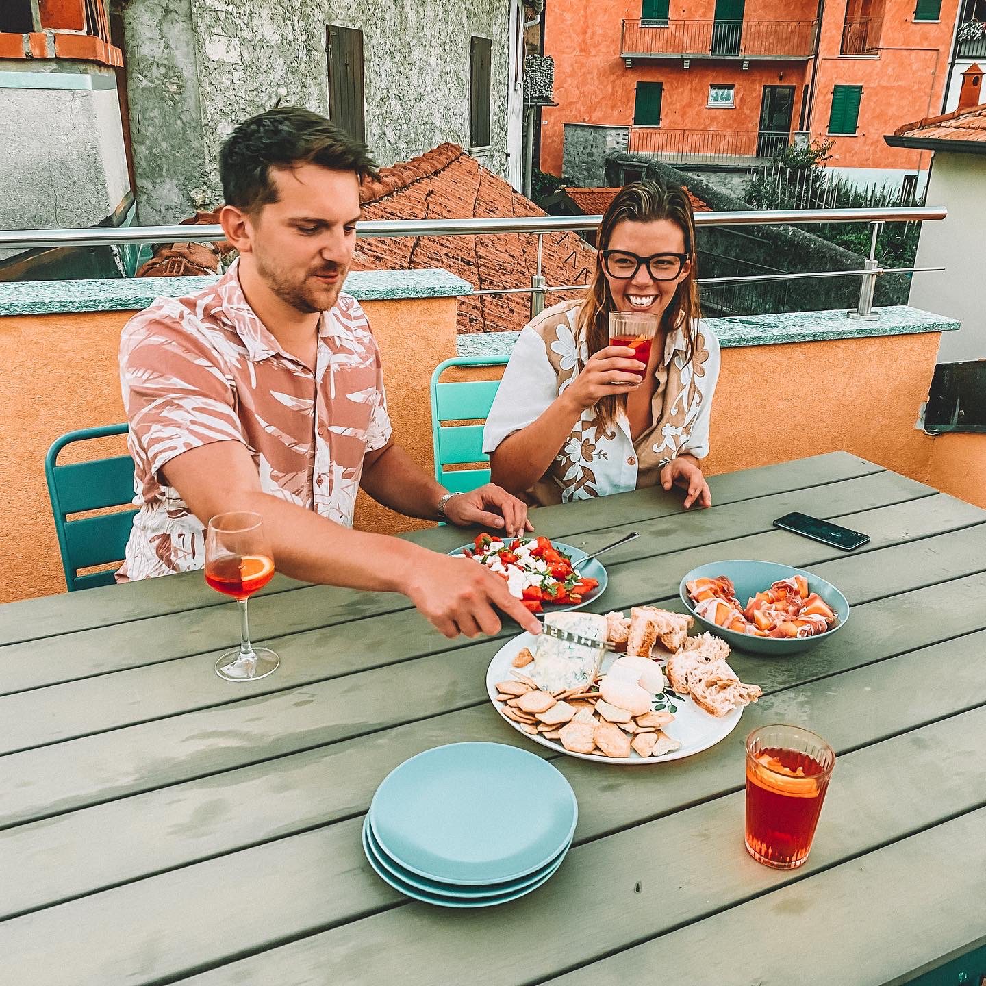 Two people drinking campari spritz on a balcony with a table in the foreground full of cheese, bread, salads and melon.