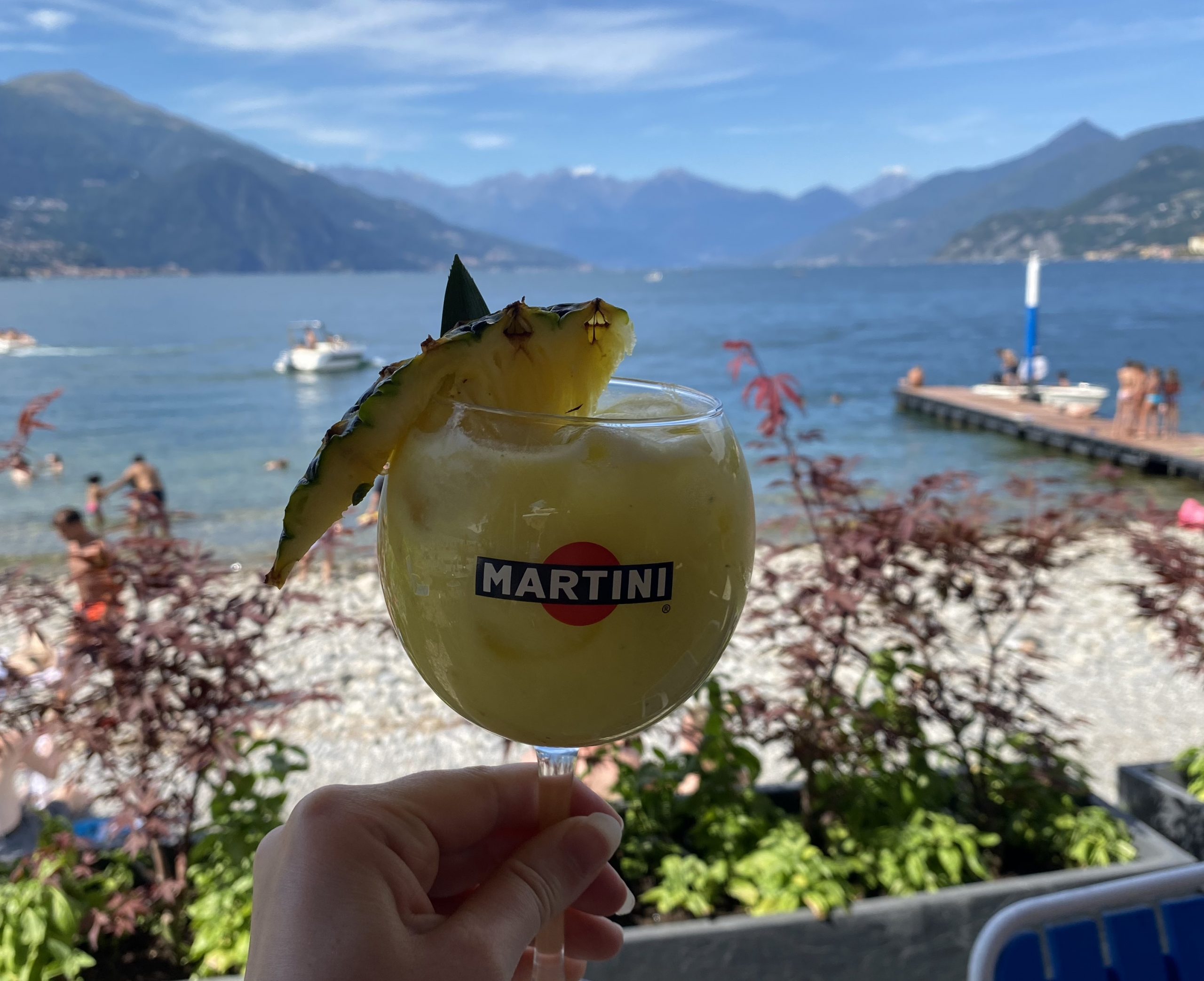 A hand holding up a yellow drink in a goblet glass with a pineapple chunk in it in front of a pebbled beach with blue water and mountains in the backdrop
