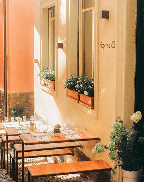 A white washed building with terracotta planters in the window and two dining tables laid with glasses and cutlery outside on the street.