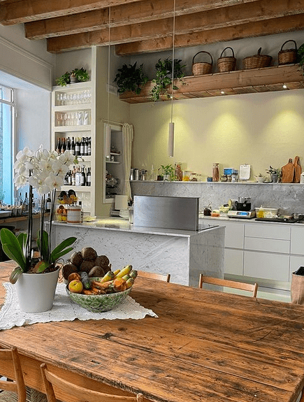 An open plan kitchen in marble and grey with bottles of wine, baskets, plants and wooden chopping boards, with a wooden table with a fruit bowl and a flowering plant in the foreground