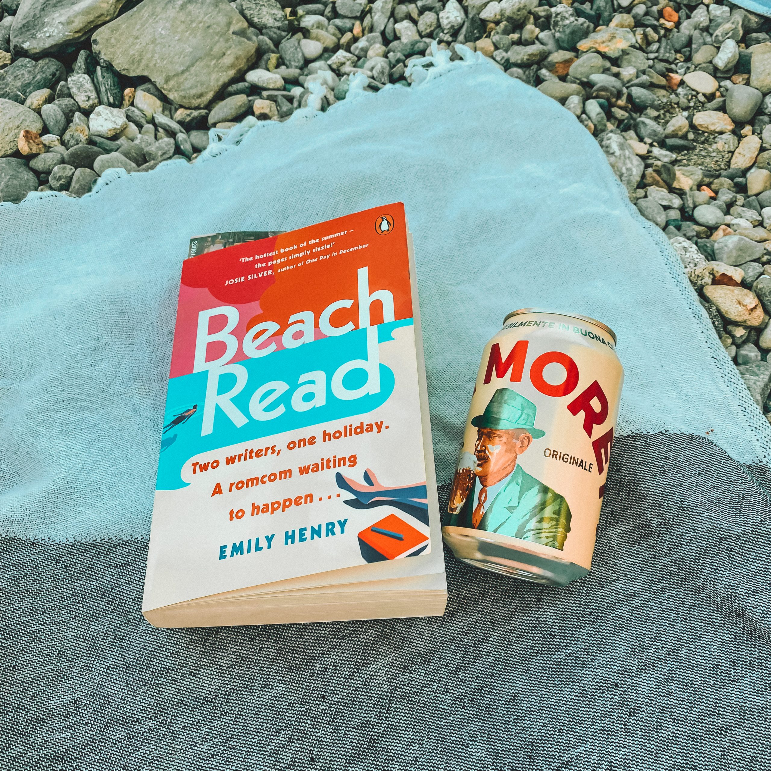 A book and a can of moretti beer on a blue beach towel on a pebble beach.