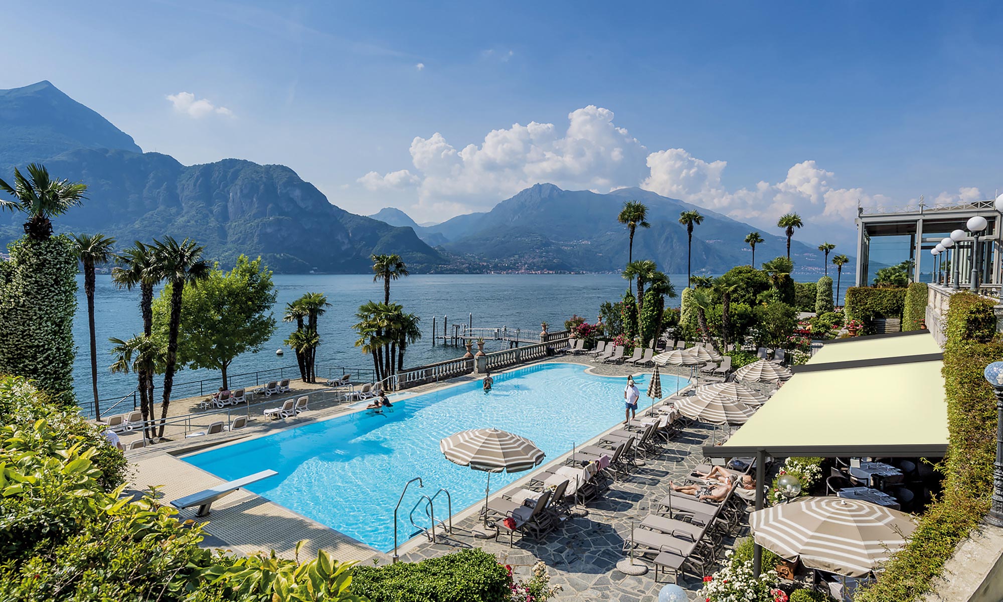 A pool lined with sun loungers, umbrellas and palm trees in front of a lake with mountains in the background