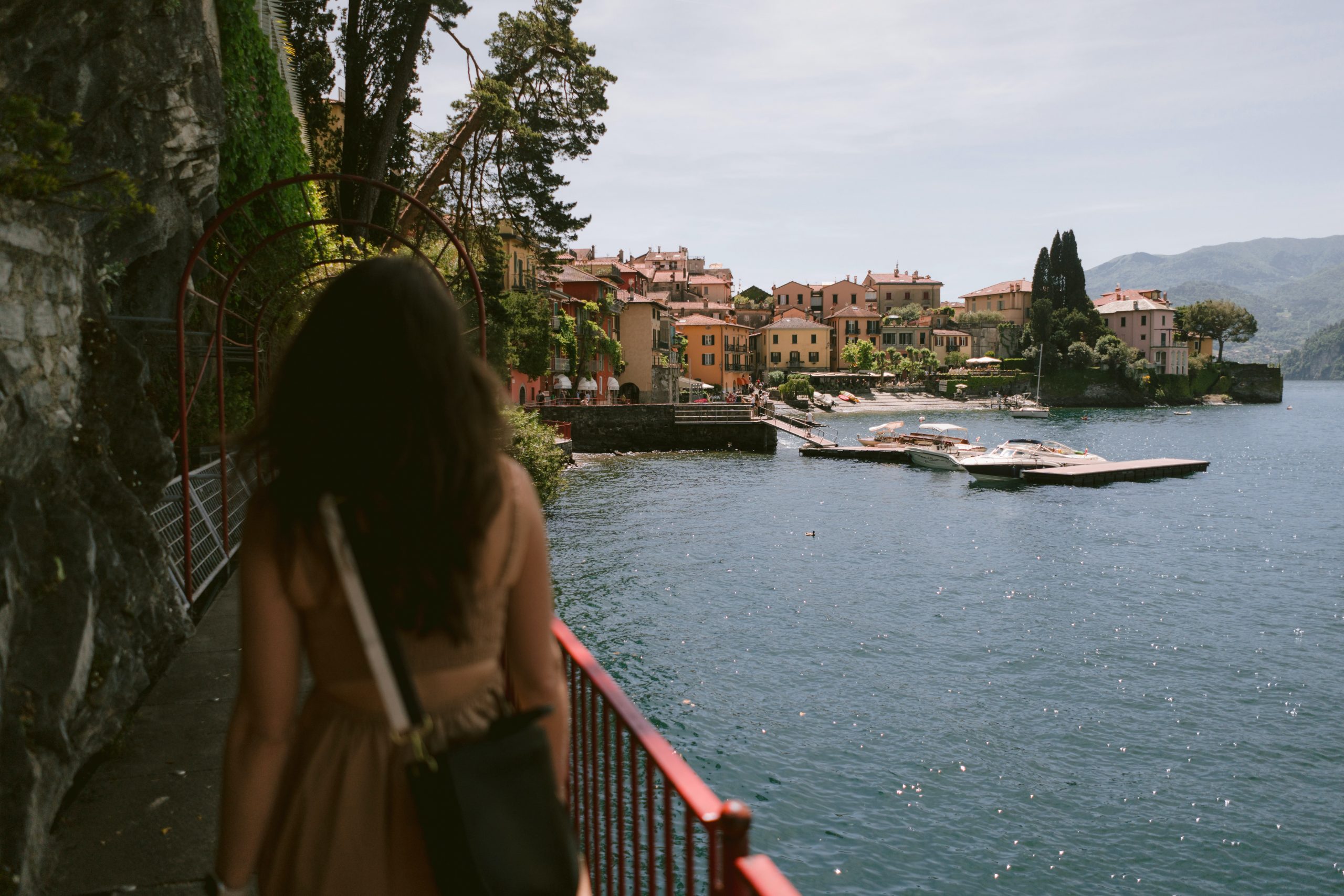 A women in a brown strapless dress walking on a lakeside walkway with views of an Italian town ahead.