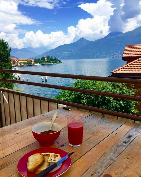 a bowl of cereal, a plate of toast and a smoothie on a wooden table overlooking a lake.