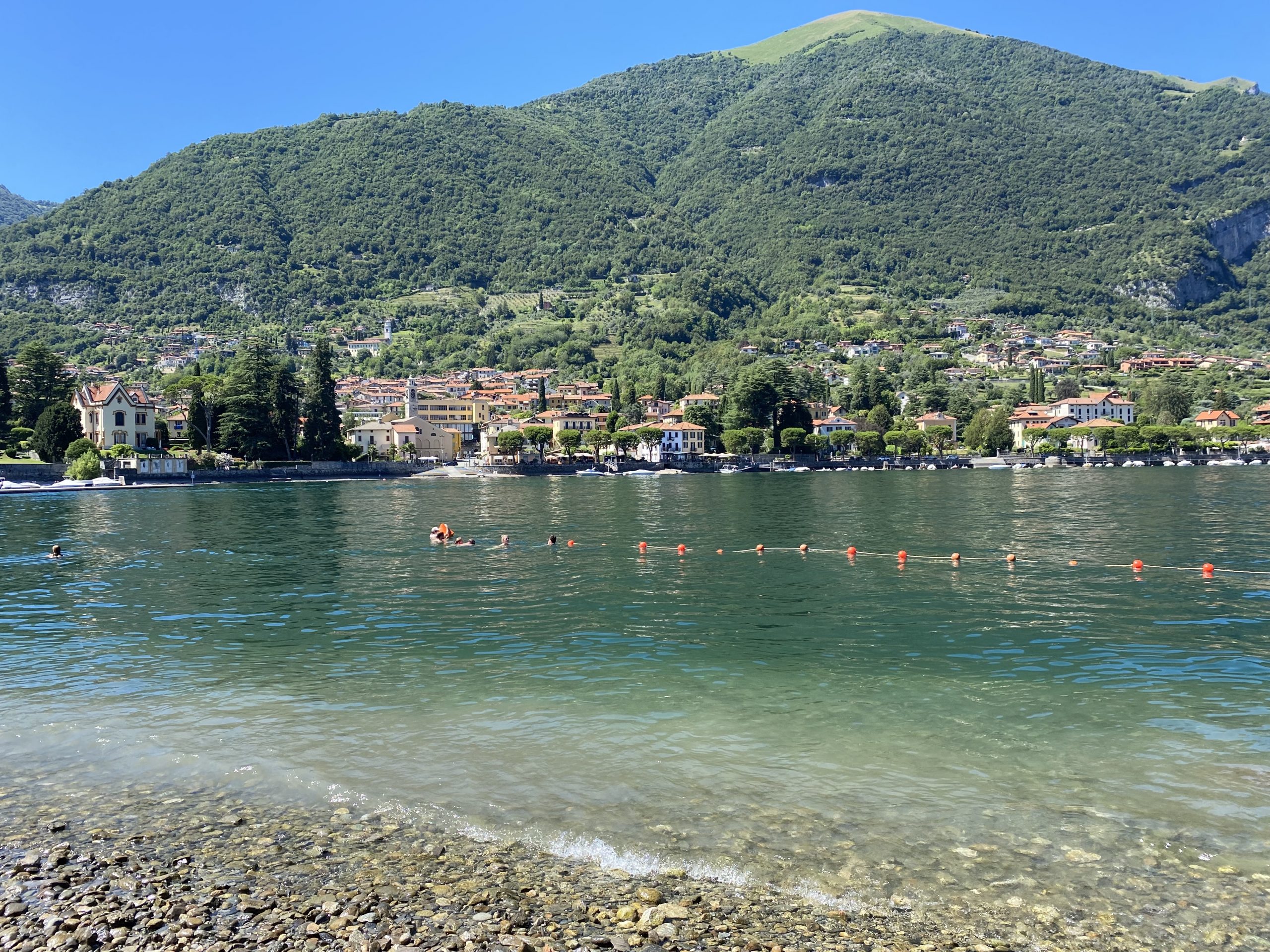 A pebble beach with orange floating buoys in the blue water and people splashing around. An Italian town with orange buildings is behind the lake set below green grassy mountains.