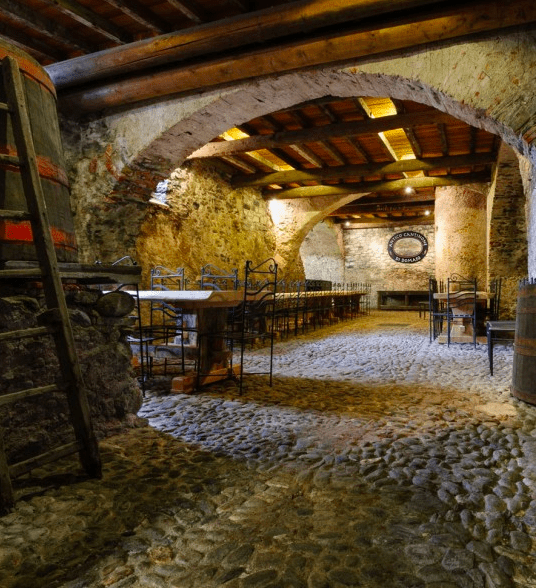 A cave style room with stone floor, archways, wooden tables and iron chairs and an old wine barrel with a ladder leading up to it.