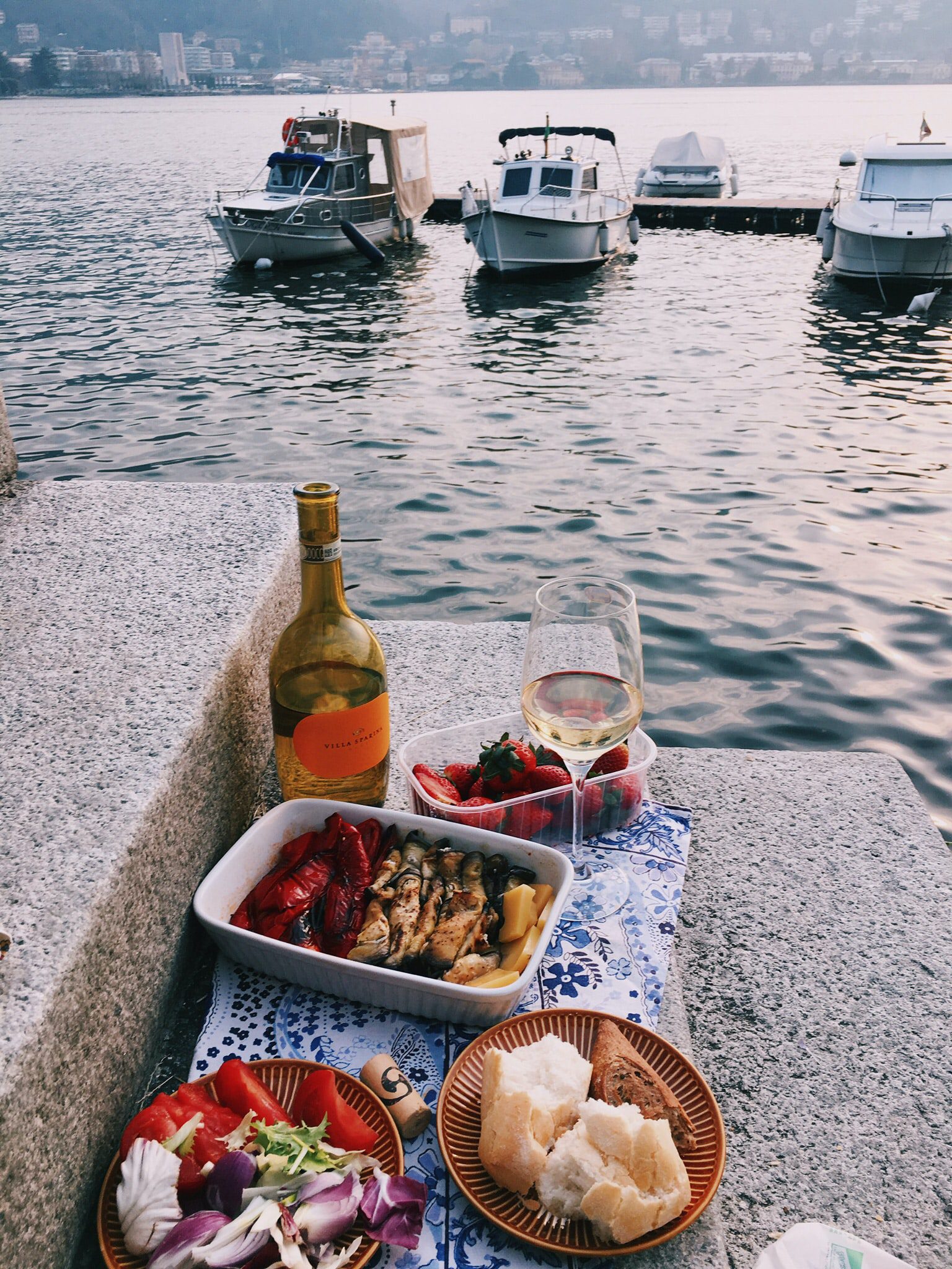 A picnic of roasted vegetables, bread, salad, strawberries and a bottle of white wine laid out on some steps overlooking a lake.