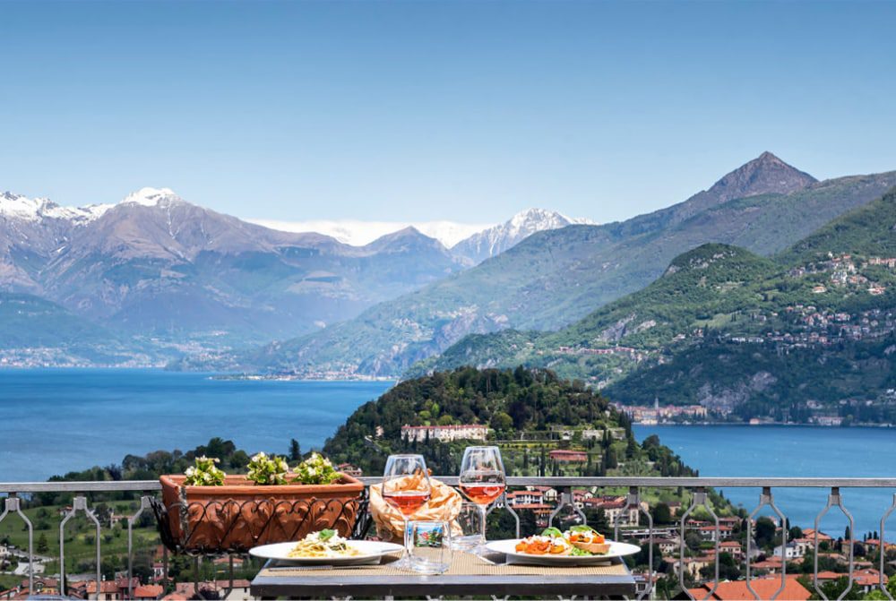 a table with plates of food and pink wine on a terrace overlooking a town below with lake and mountains in the background