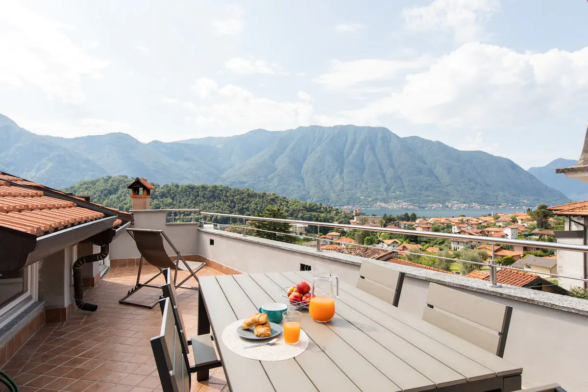a jug of orange juice, pastries and fruit on top of a table on a rooftop overlooking a lake and mountains.