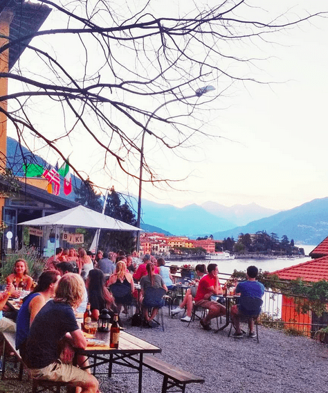 people clustered around tables on a gravelled outdoor terrace overlooking a lake.