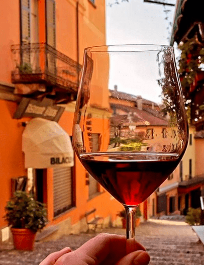 a wine glass filled with red liquid being held up by a hand in front of a cobbled street with orange buildings