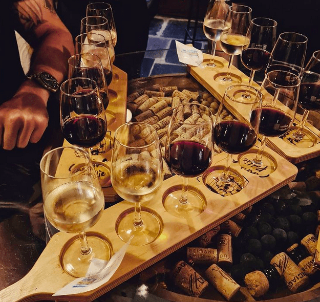 A selection of wine glasses half full on wooden paddles on top of a big wine barrel which has corks stuck on top of it underneath a glass sheet.