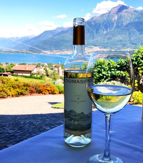 A bottle of white wine and a glass half full set on a white clothed table overlooking a blue lake and mountains in the distance.