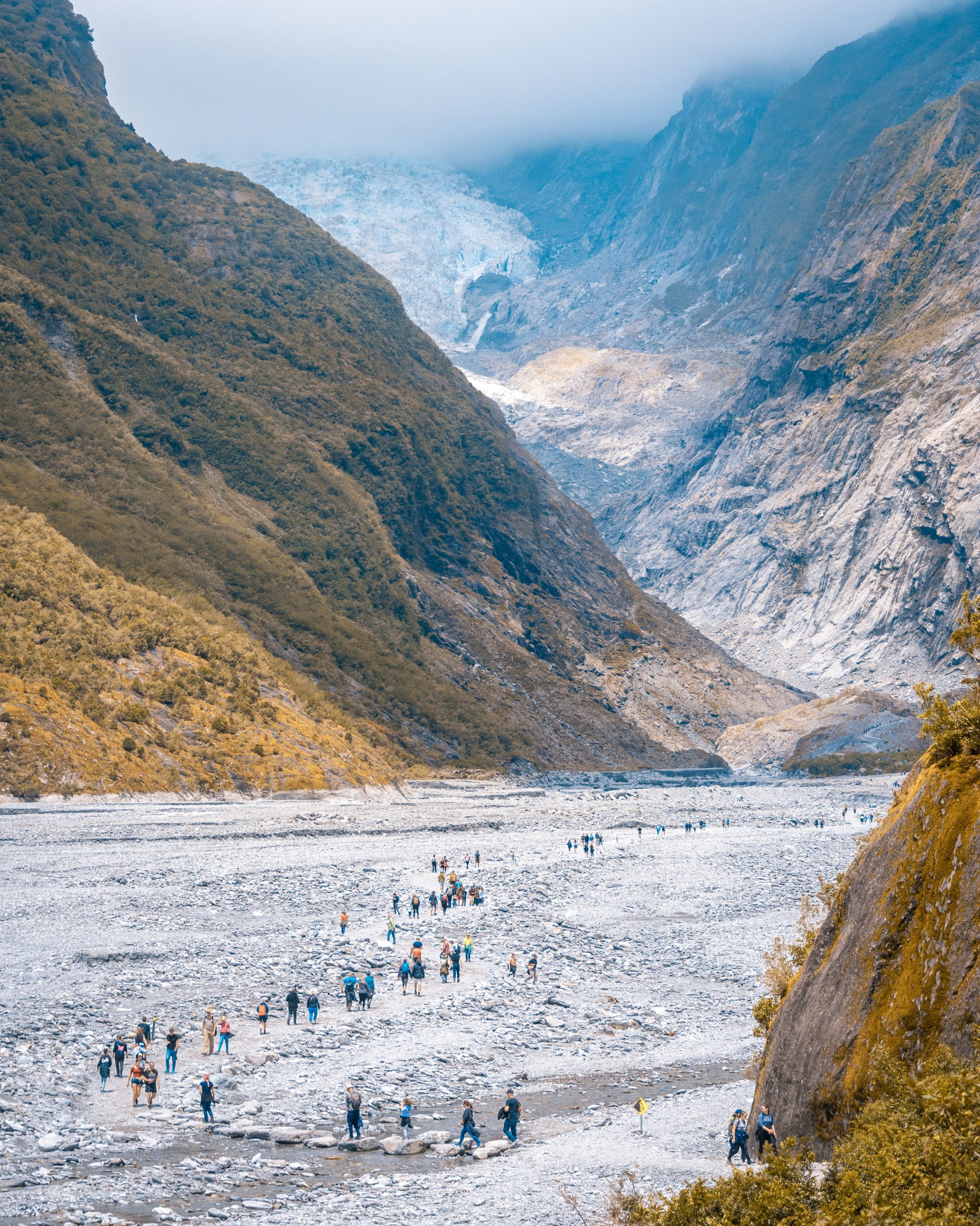 A retreating icy glacier with a rocky river bed in the foreground used by people walking towards the glacier.