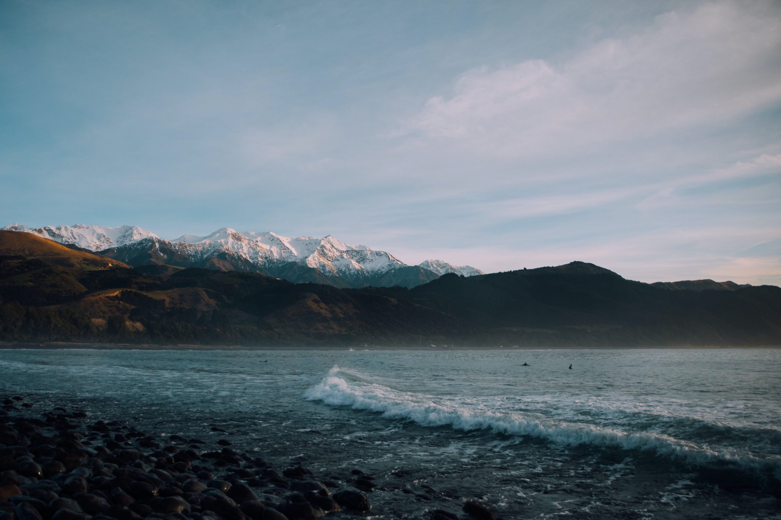 A rocky beach with gentle waves surrounded by snow-capped mountains.