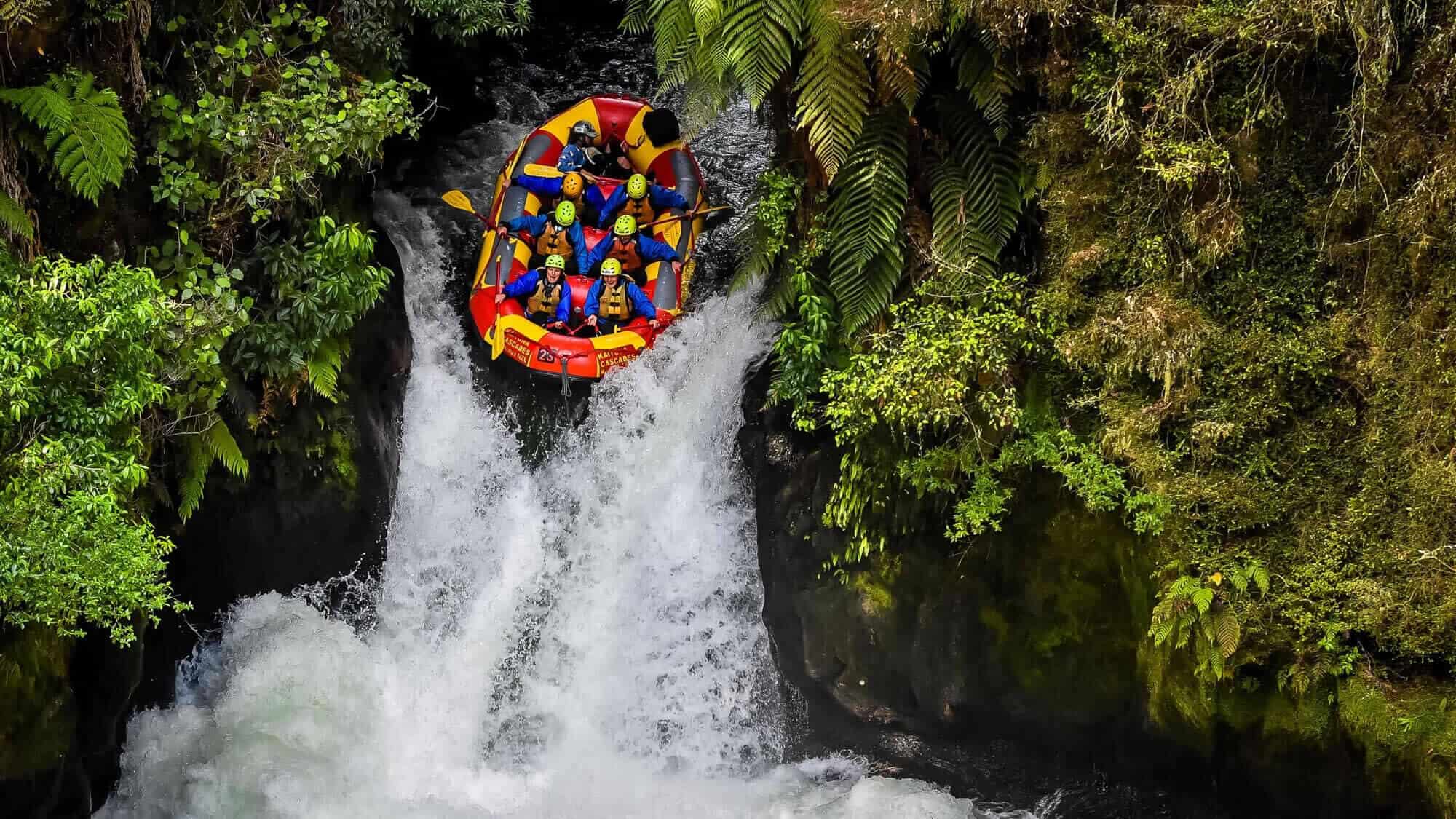 An inflatable raft filled with people dropping down a steep waterfall with green lush plants and rocks on either side.