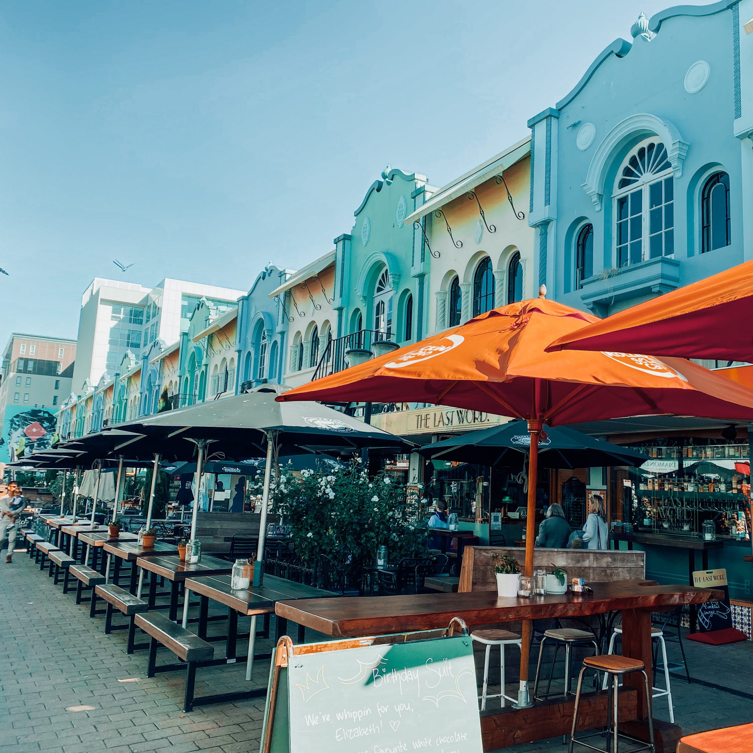 A street lined with colourful regent-style buildings with outdoor tables, benches and parasols out front.