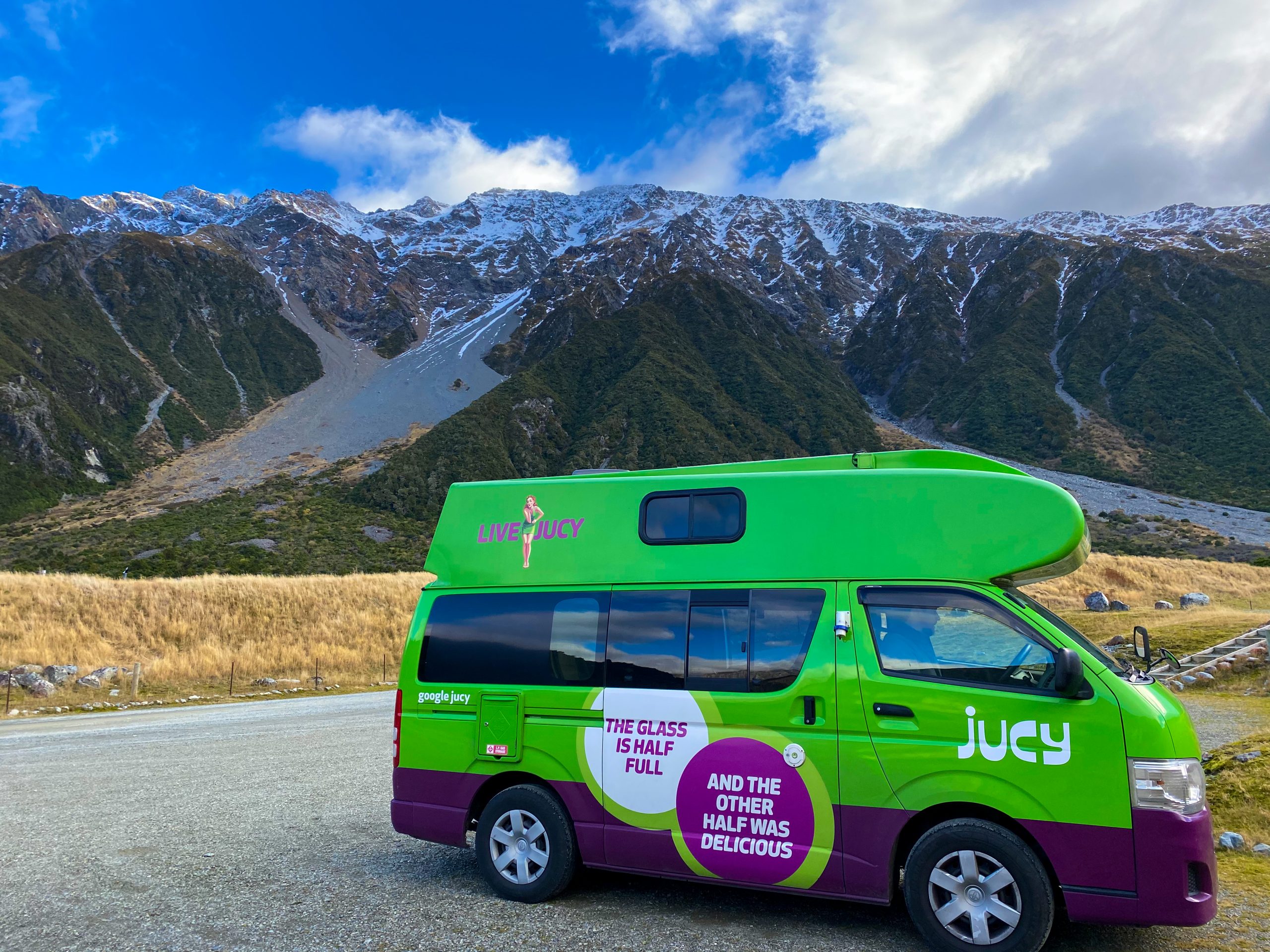 A green and purple campervan in a grassy camping spot overlooking snow-capped mountains.