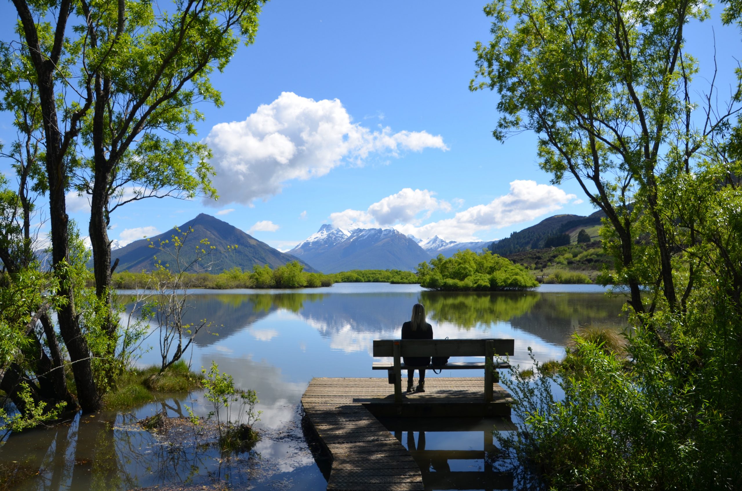 A person sitting on a bench on a wooden boardwalk jutting out onto a lake with trees and mountains in the distance.