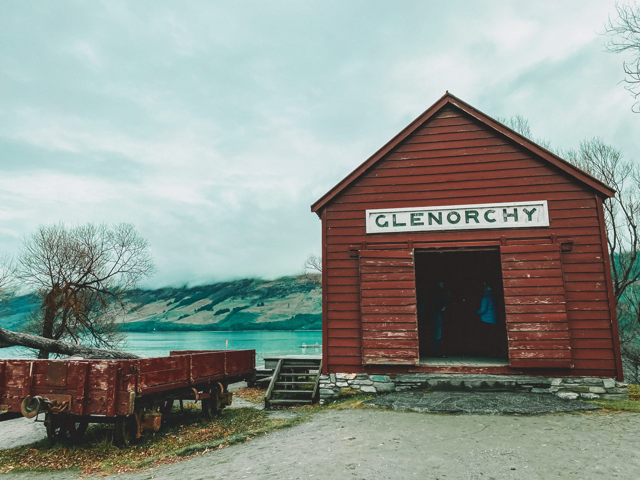 An old red boat hut with a white sign reading 'GLENORCHY' at the top and an old red carriage to the left, in front of a lake and mountains covered in fog.