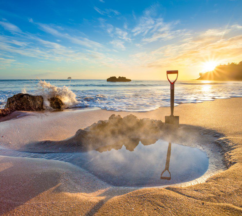 A steaming pool of water on a beach with a shovel stuck in the sand. Waves crash on rocks and the sun rises in the distance.