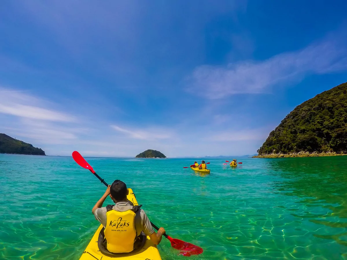 People kayaking on yellow kayaks with red paddles on a body of clear blue water with islands and lush peninsulas in the distance.