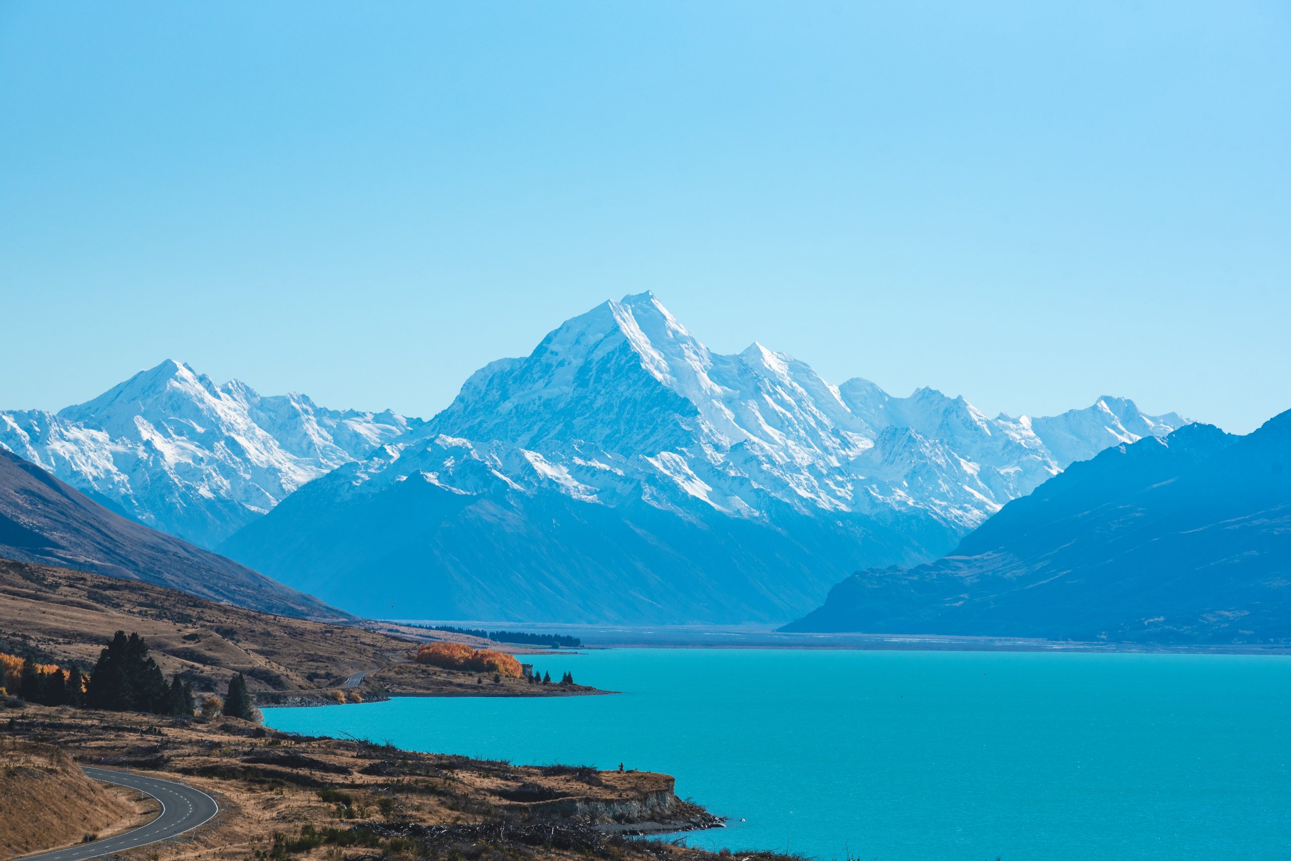 A turquoise blue lake in front of towering snow-capped mountains with a winding road on the left leading towards the mountain range.