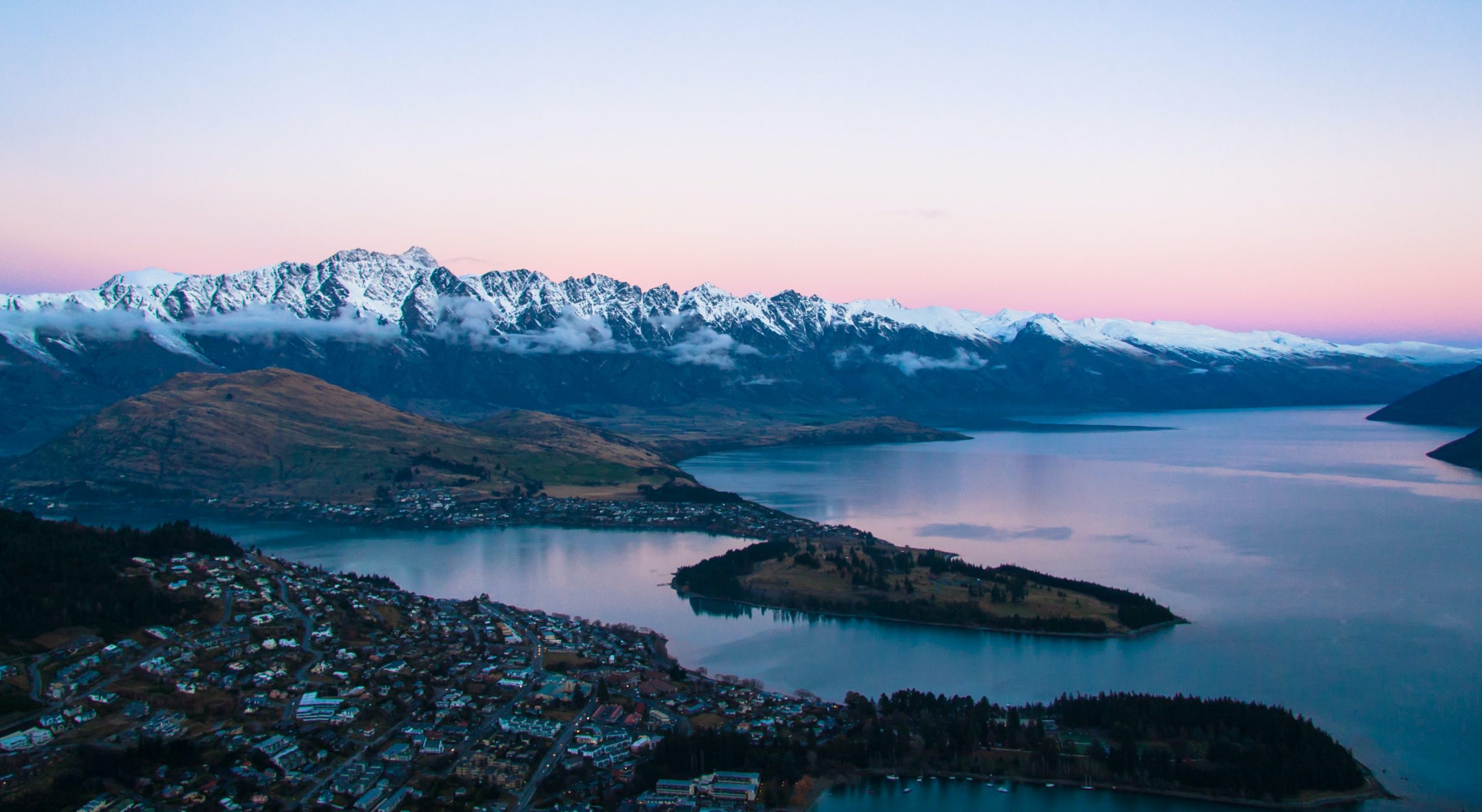 An aerial view of a lakeside town at sunrise overlooked by snow-capped mountains.