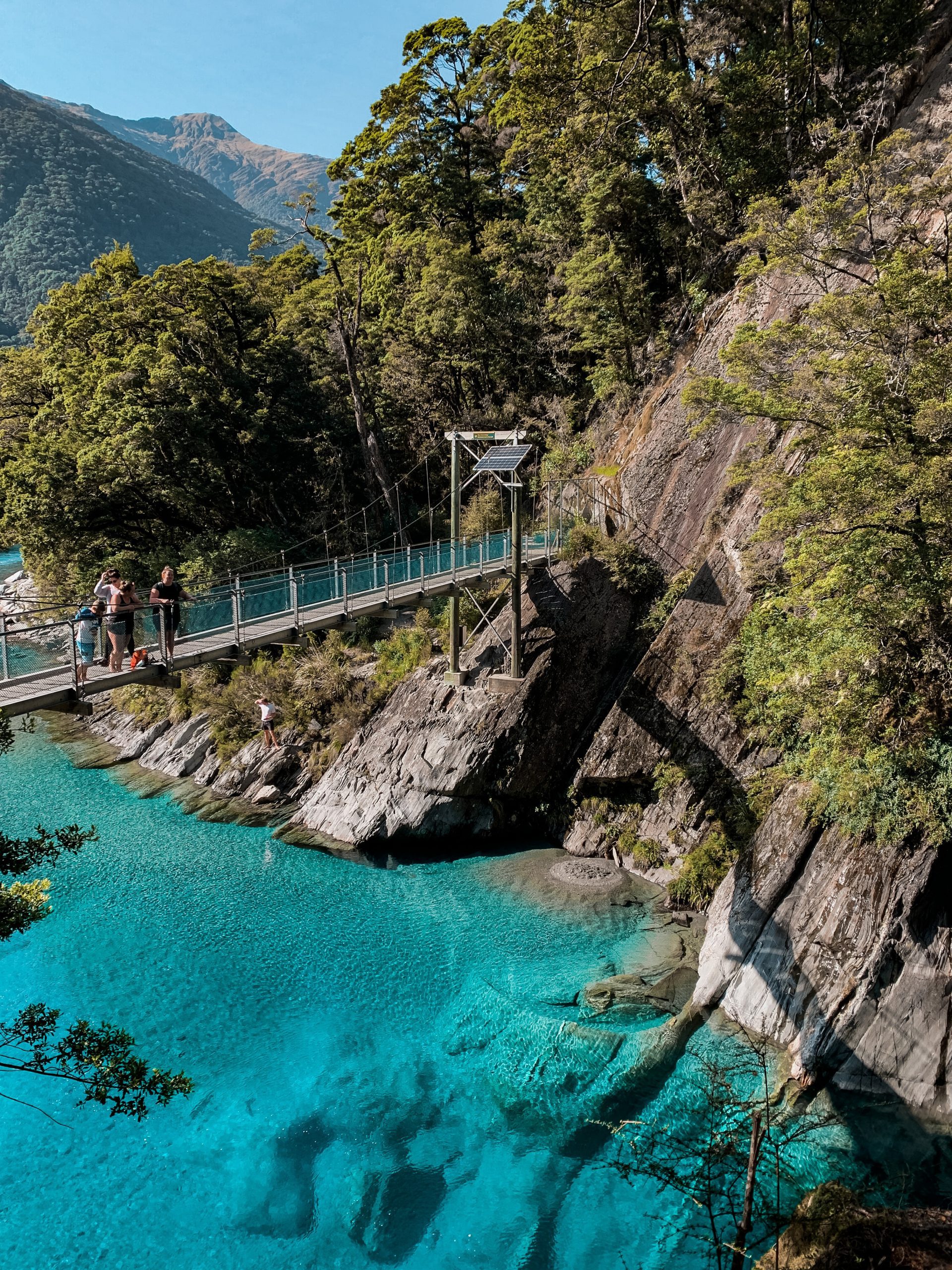 A family with children standing on a swing-bridge above bright turquoise water and cliffs/ trees in the backdrop.