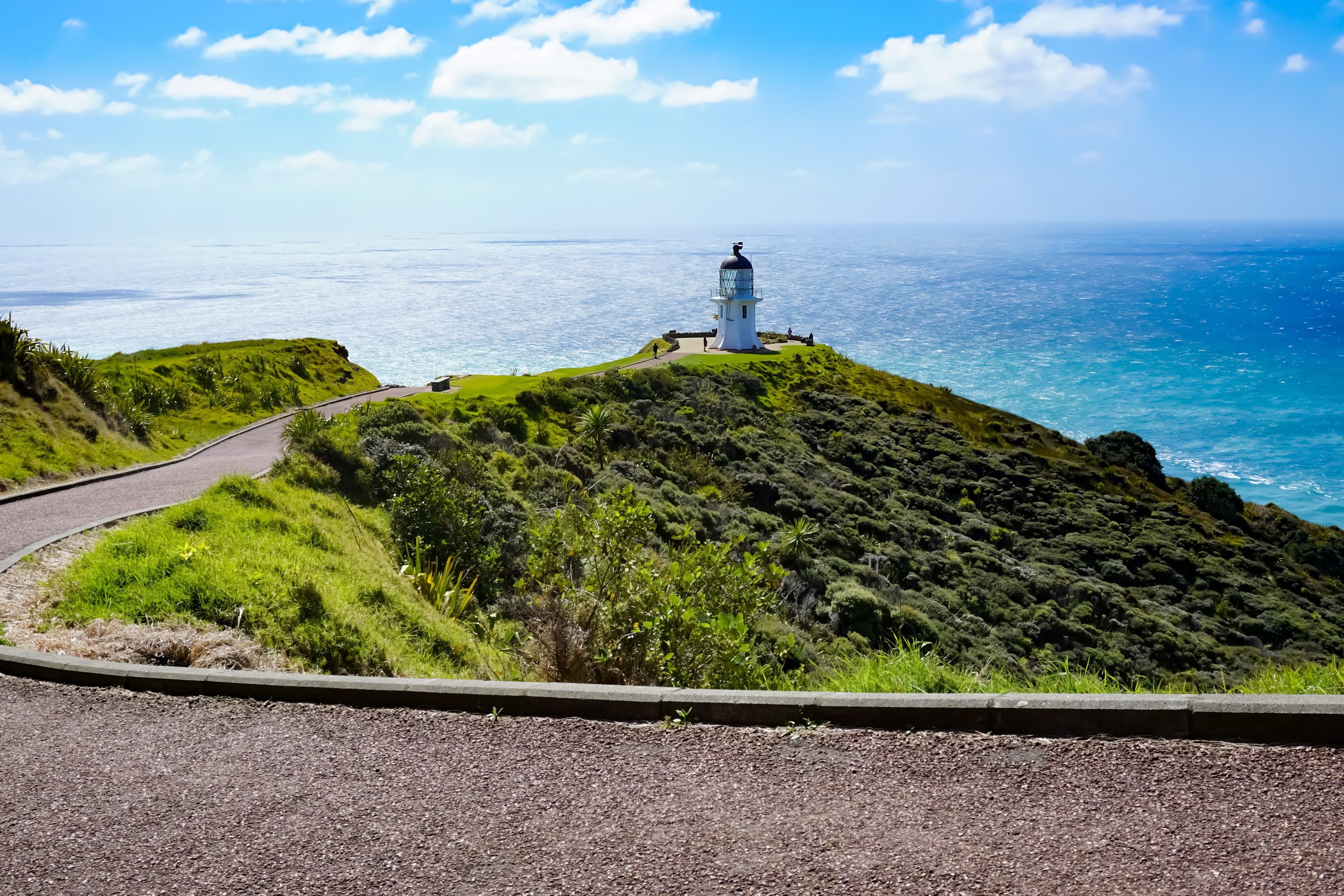 A tarmac path leading down to a small white lighthouse surrounded by grassland and the blue ocean beyond it.