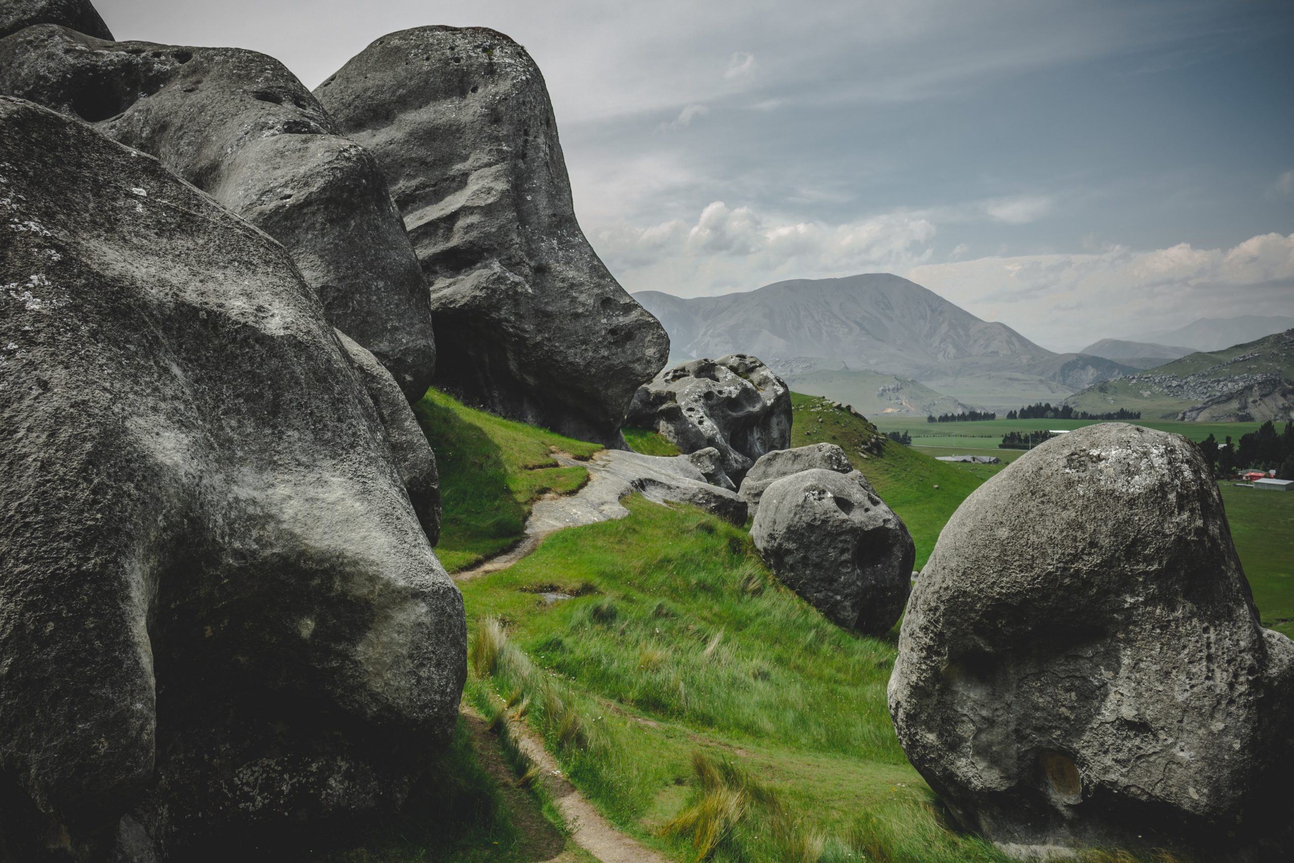 A green grassy path winding through giant grey boulders, with mountains in the backdrop.