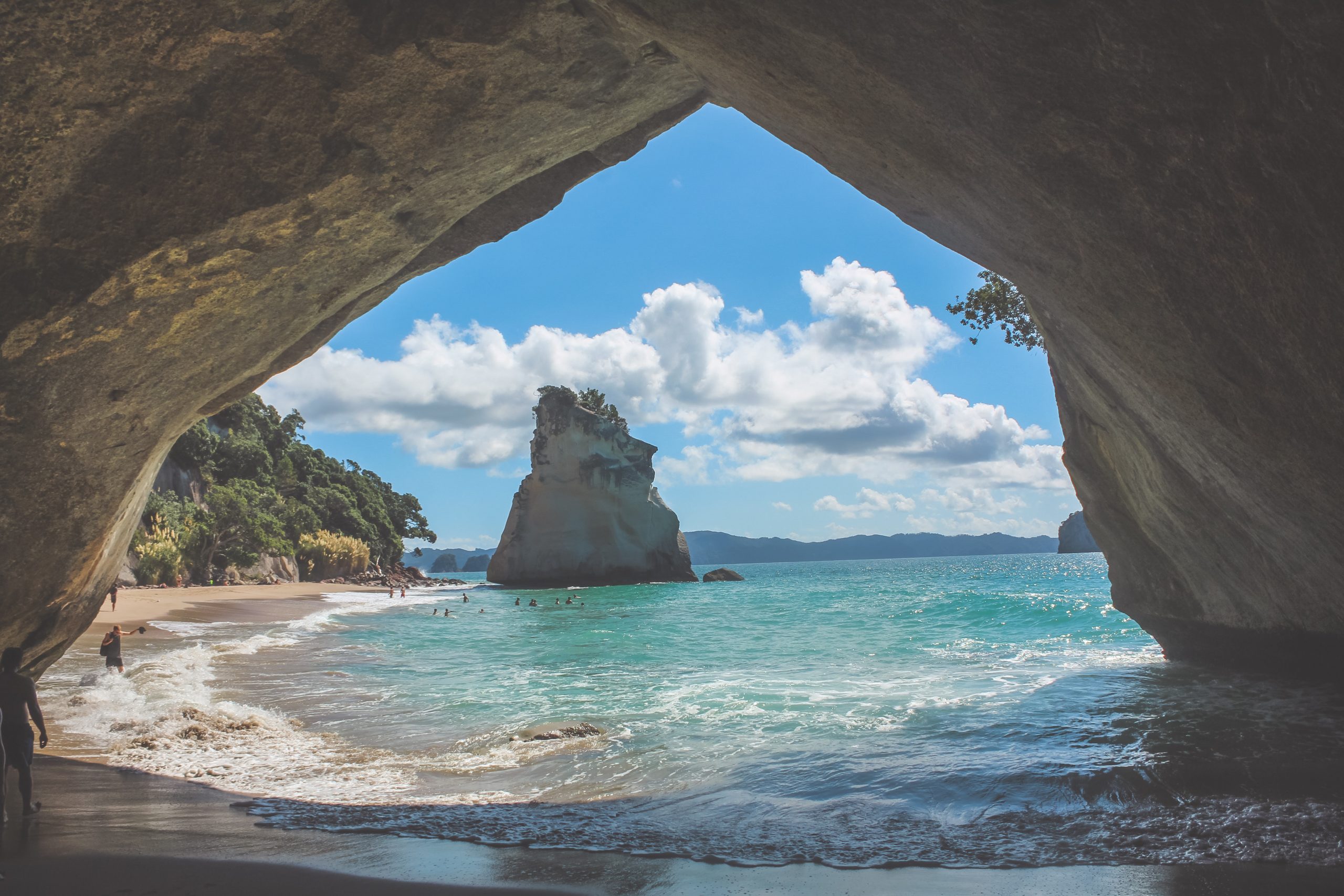 The view of a golden sandy cove with blue ocean and a large rock, seen through a natural stone archway.