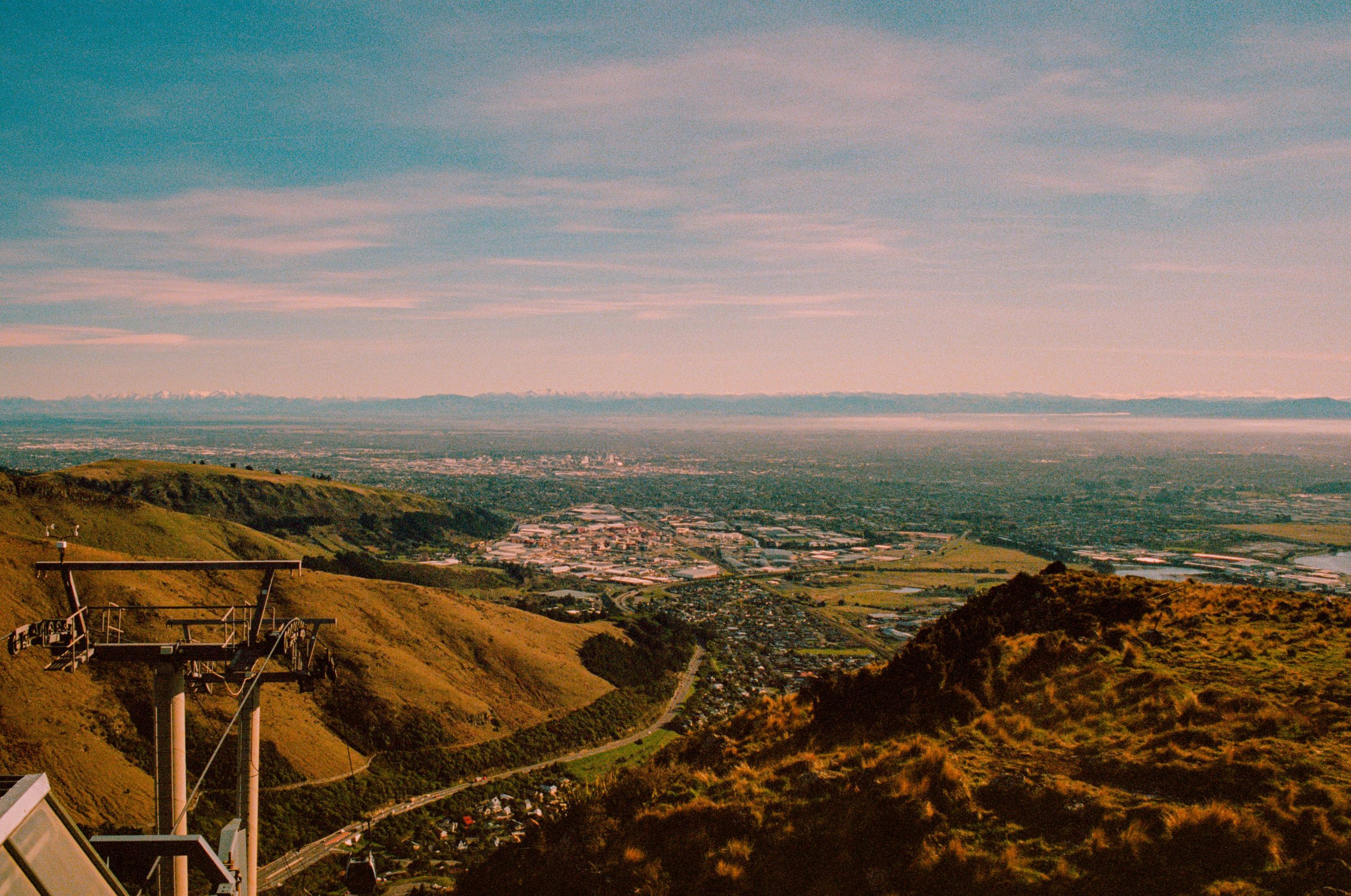 A cable car pylon at the top of a hill with grassy slopes, a city and a harbour in the background.