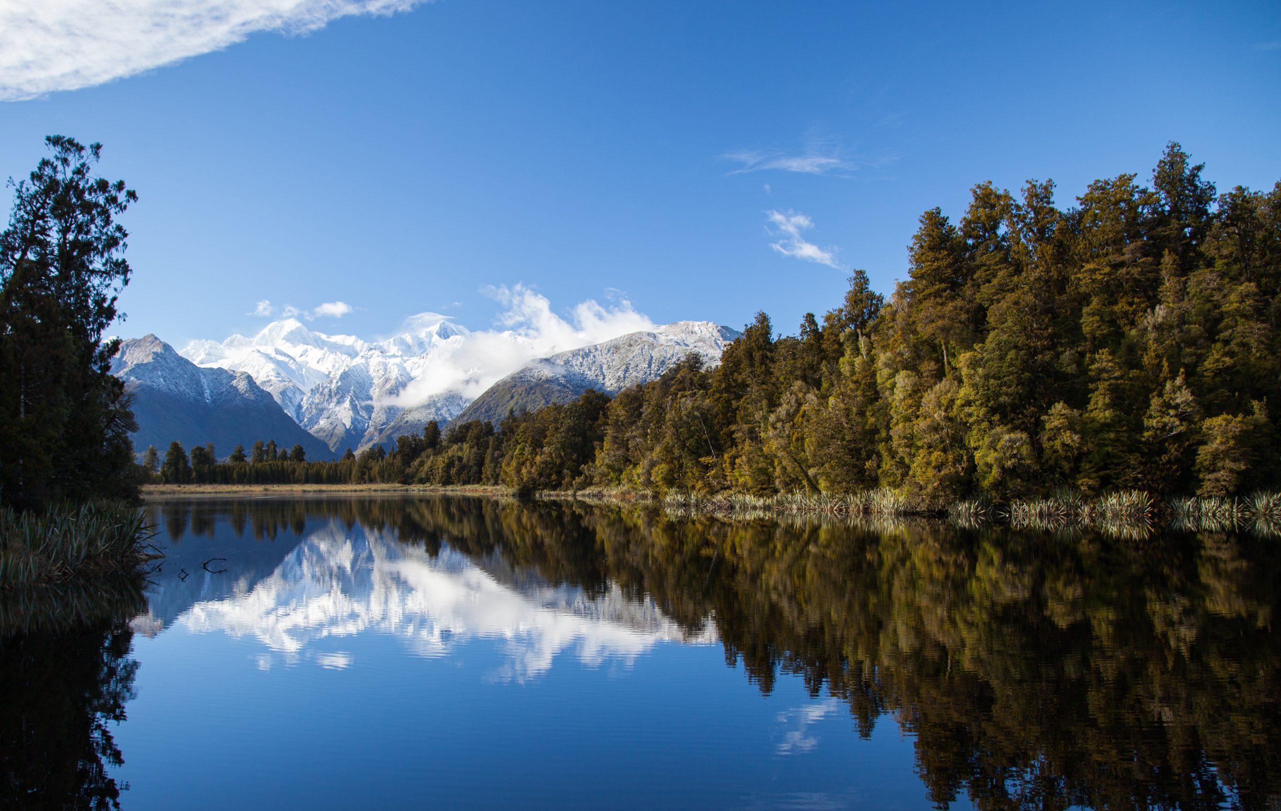 A reflective lake with snow-capped mountains reflected back into the water and forest on each side.