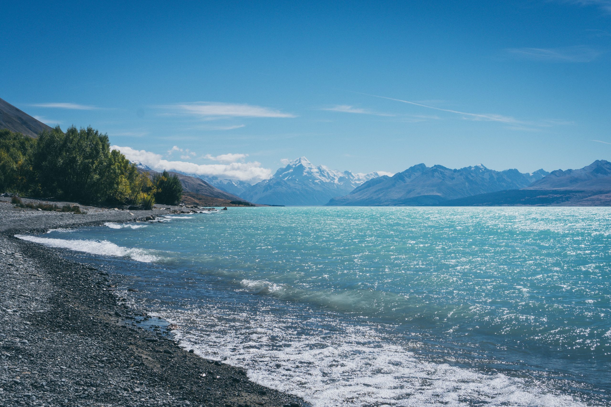 A bright turquoise lake with a black rocky shoreline flanked by snow-capped mountains in the distance.