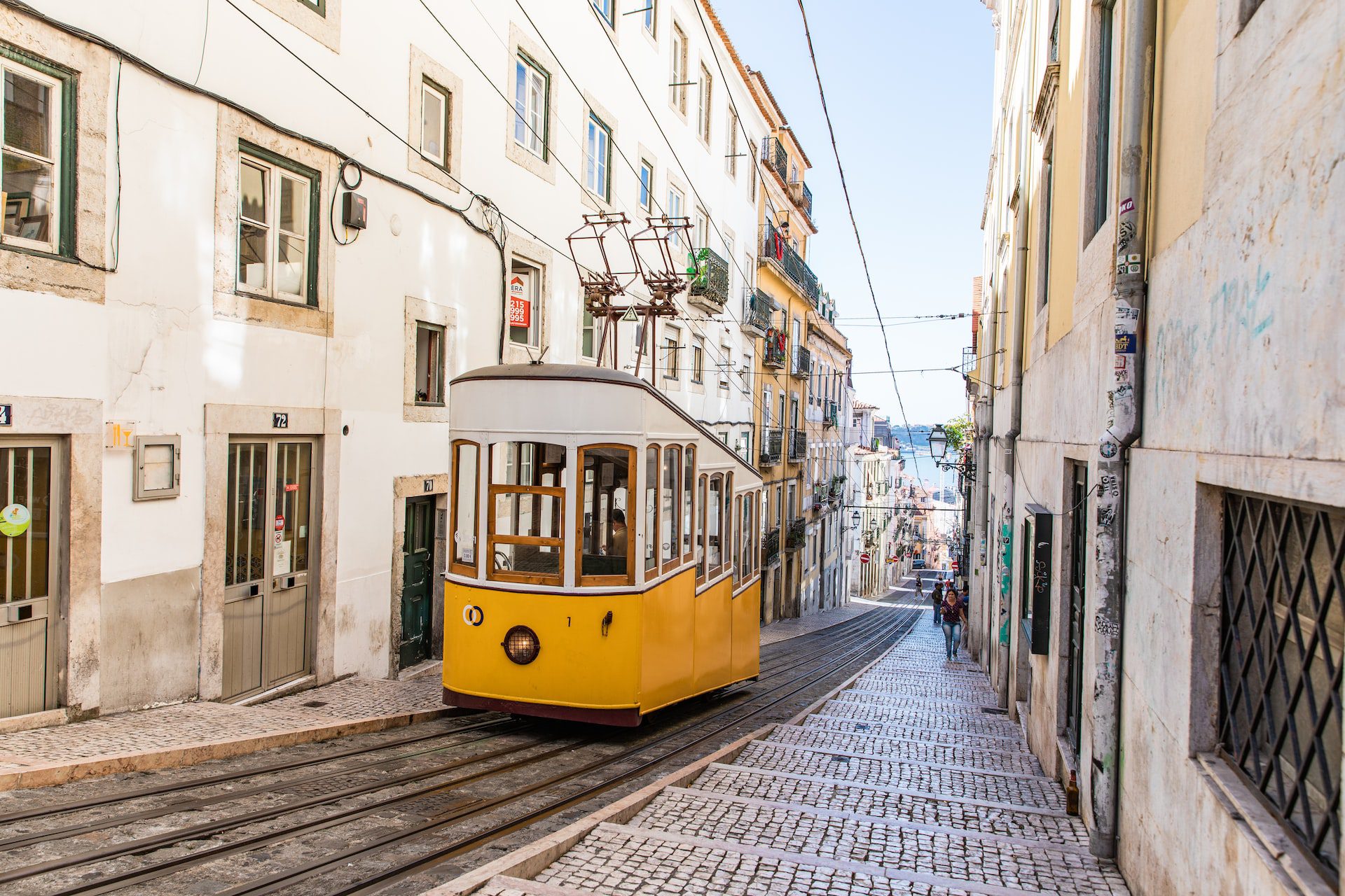 A yellow tram on a steep cobbled street lined with white and yellow buildings.