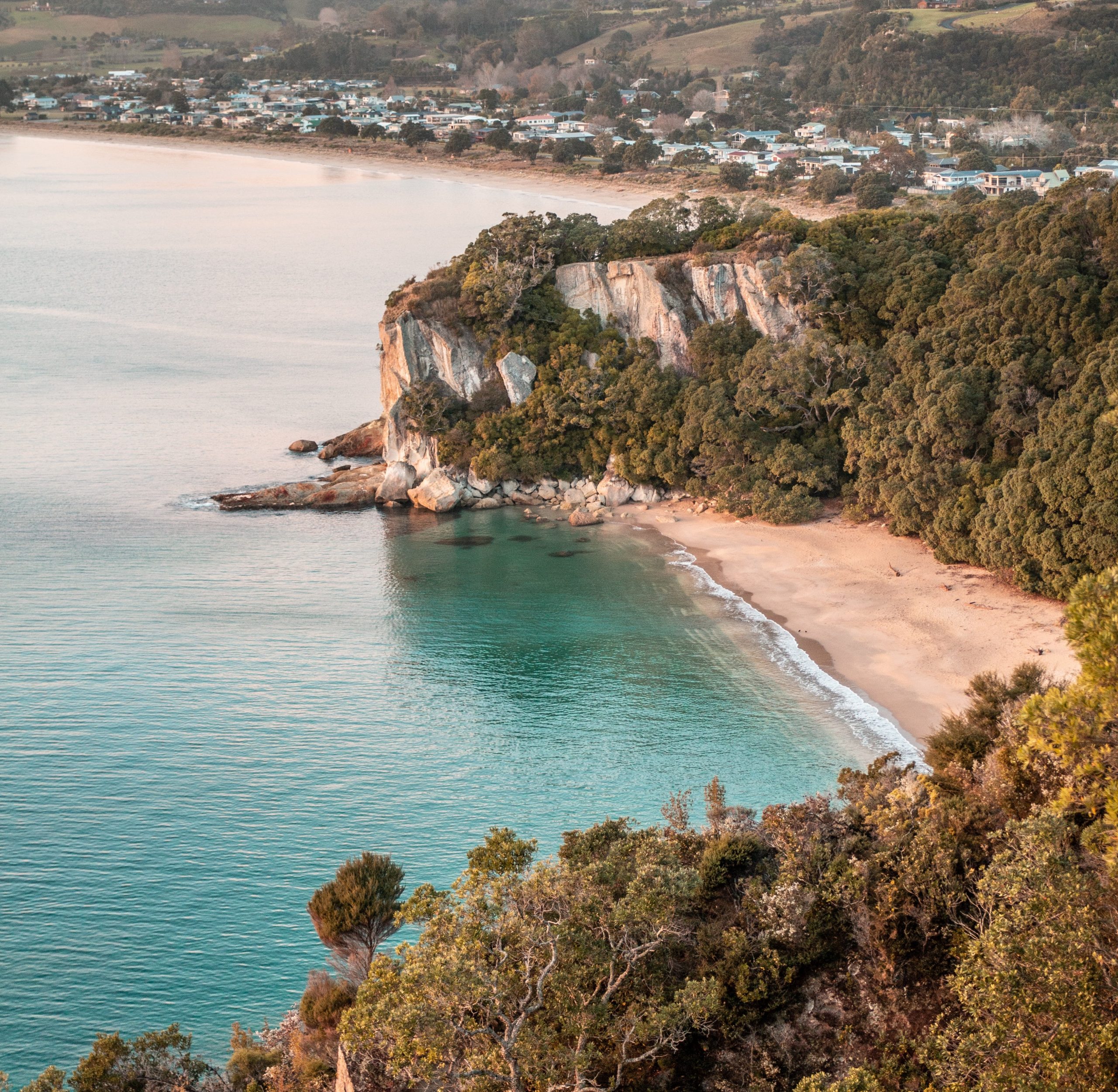 A vantage point overlooking a small bay with golden sand and gentle turquoise waves with a cliff behind it and a seaside town with houses and telephone lines.