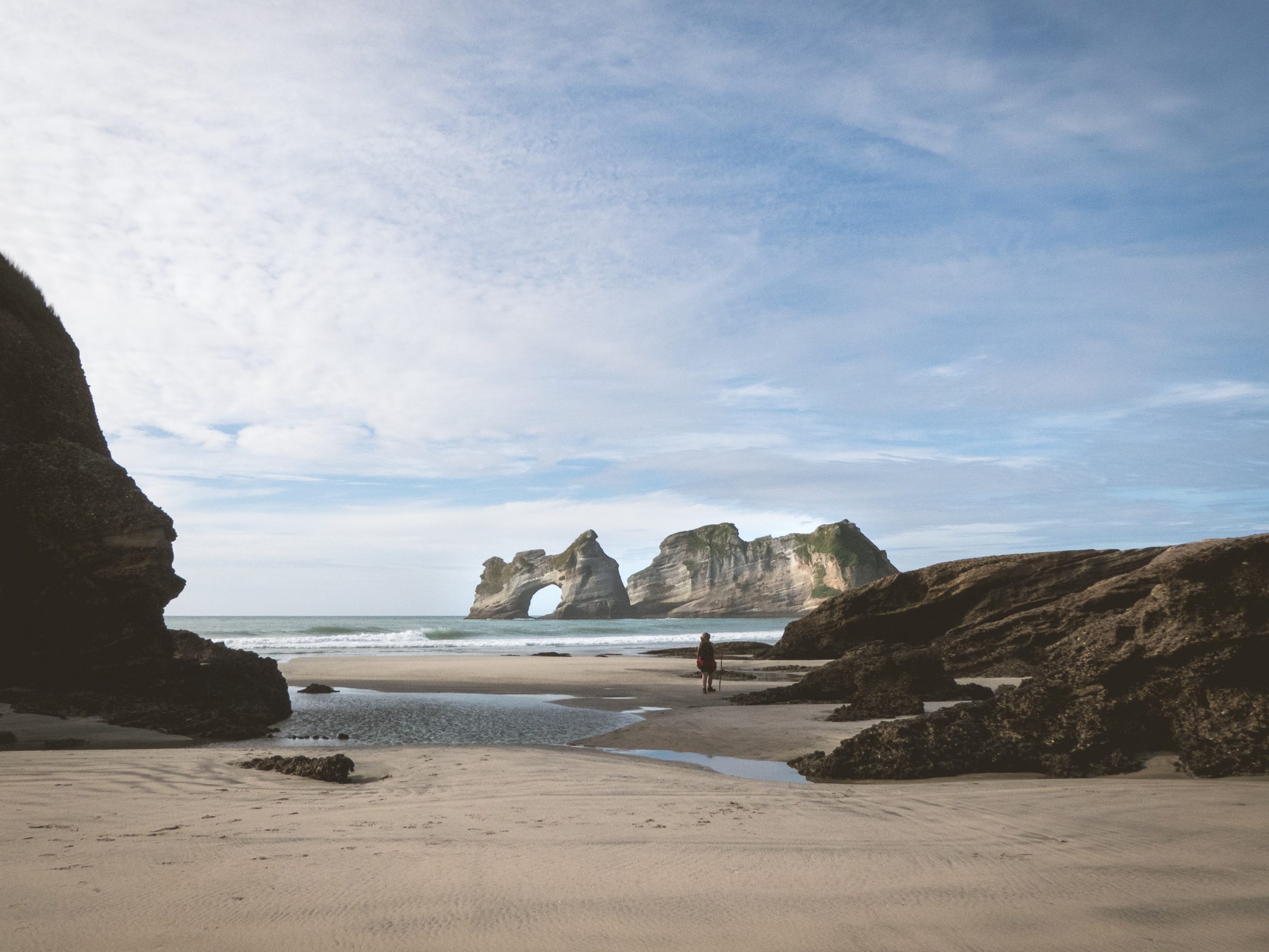 A sandy beach with large black rocks and a natural rock arch island in the ocean in the distance.