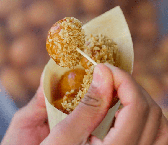 A person picking up a small round donut topped with nuts from a brown paper cone using a toothpick.