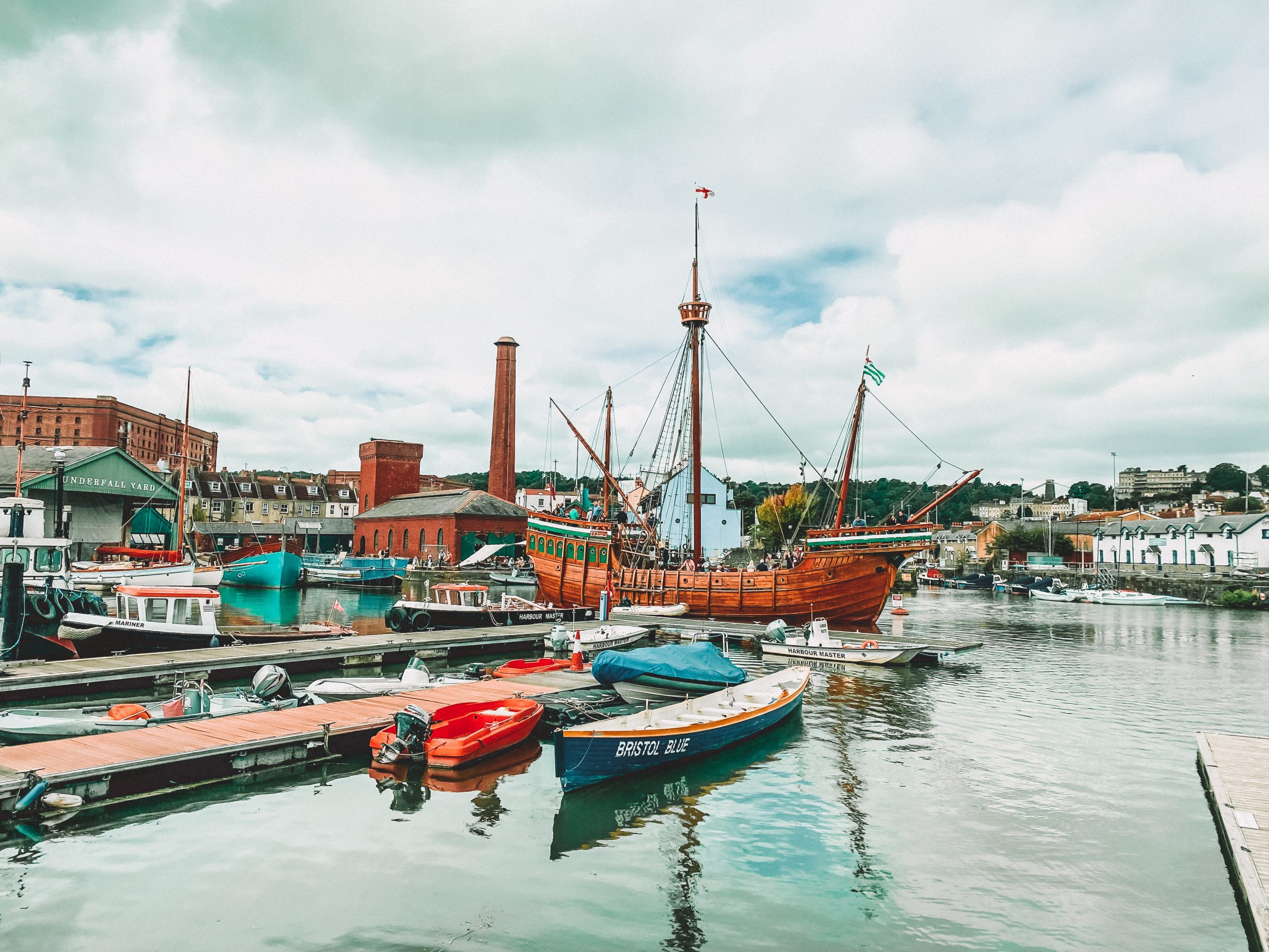 A harbour with an old wooden pirate ship, life boats and small rowing boats, with boat yards and a mill in the background.