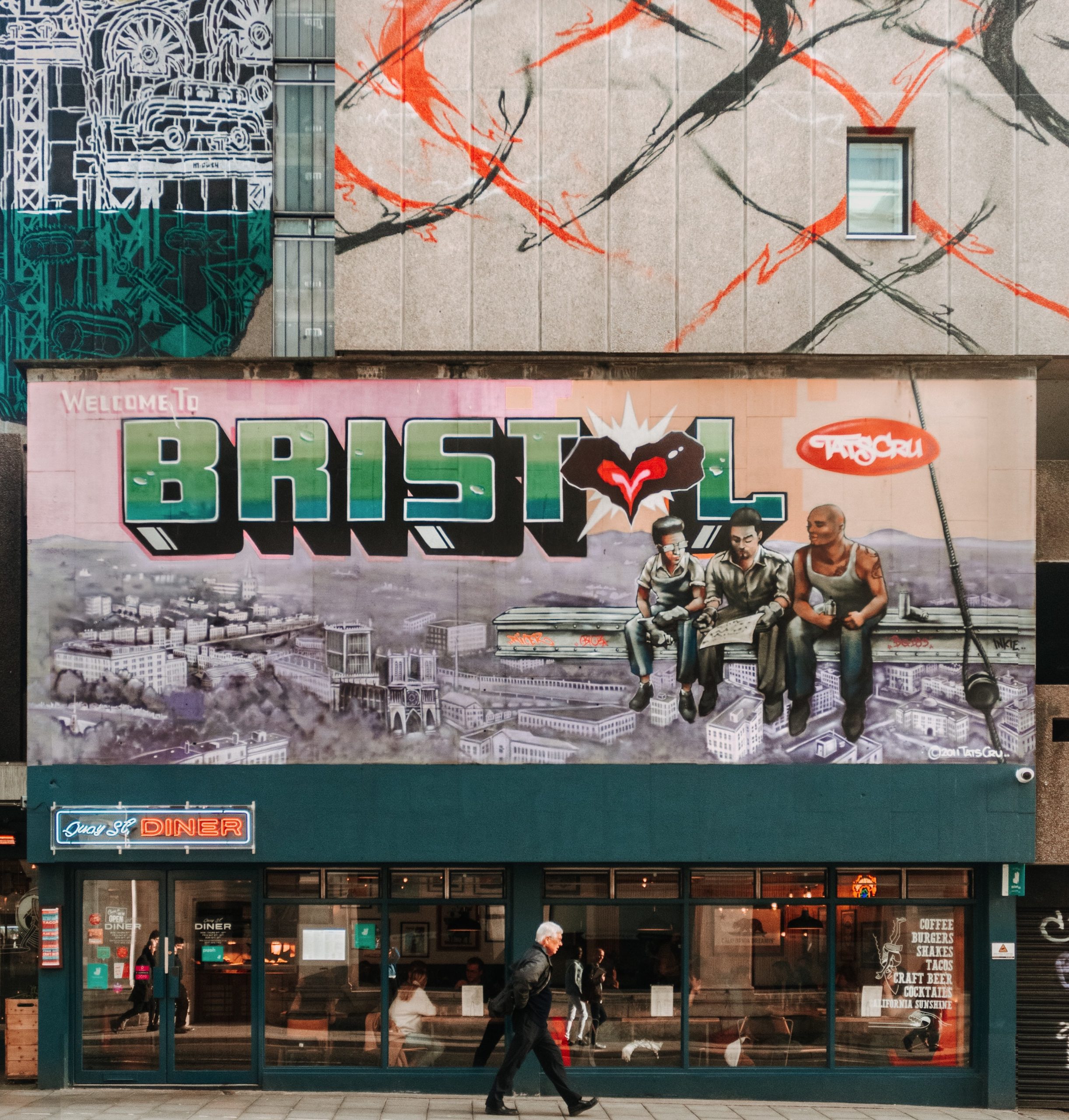 A man walking past a diner with a huge street art mural above it saying 'welcome to Bristol'.
